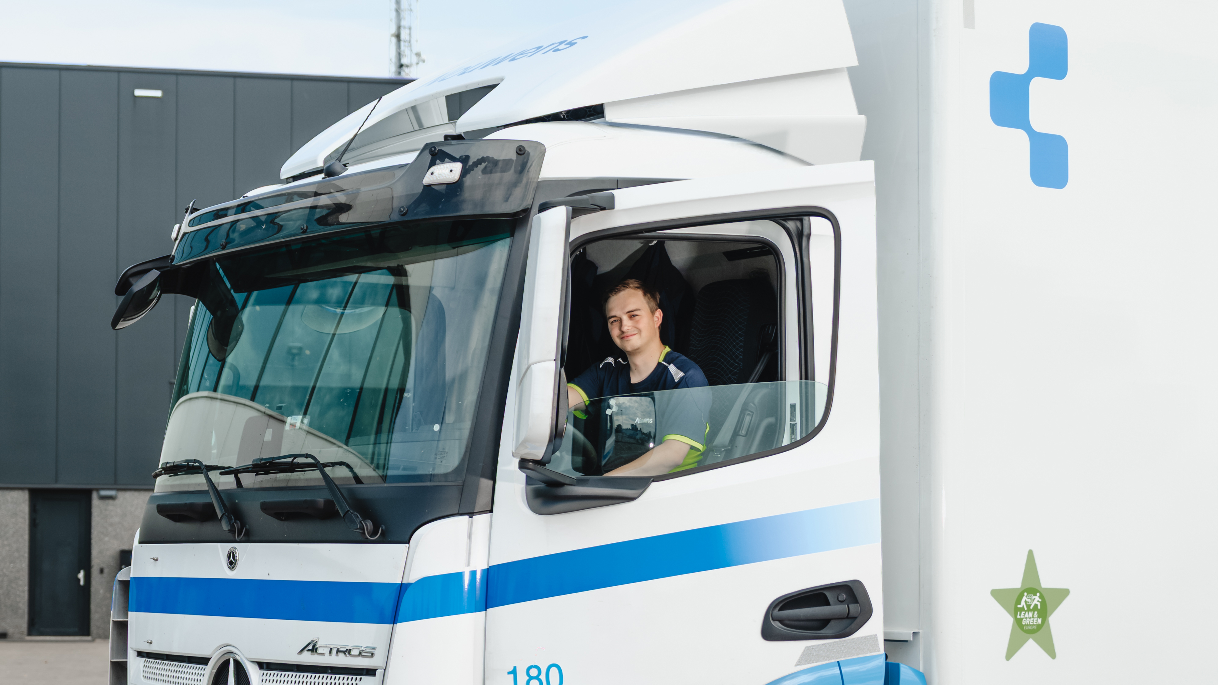 Smiling truck driver sitting in the cab of a white Mercedes-Benz Actros truck with blue stripes.