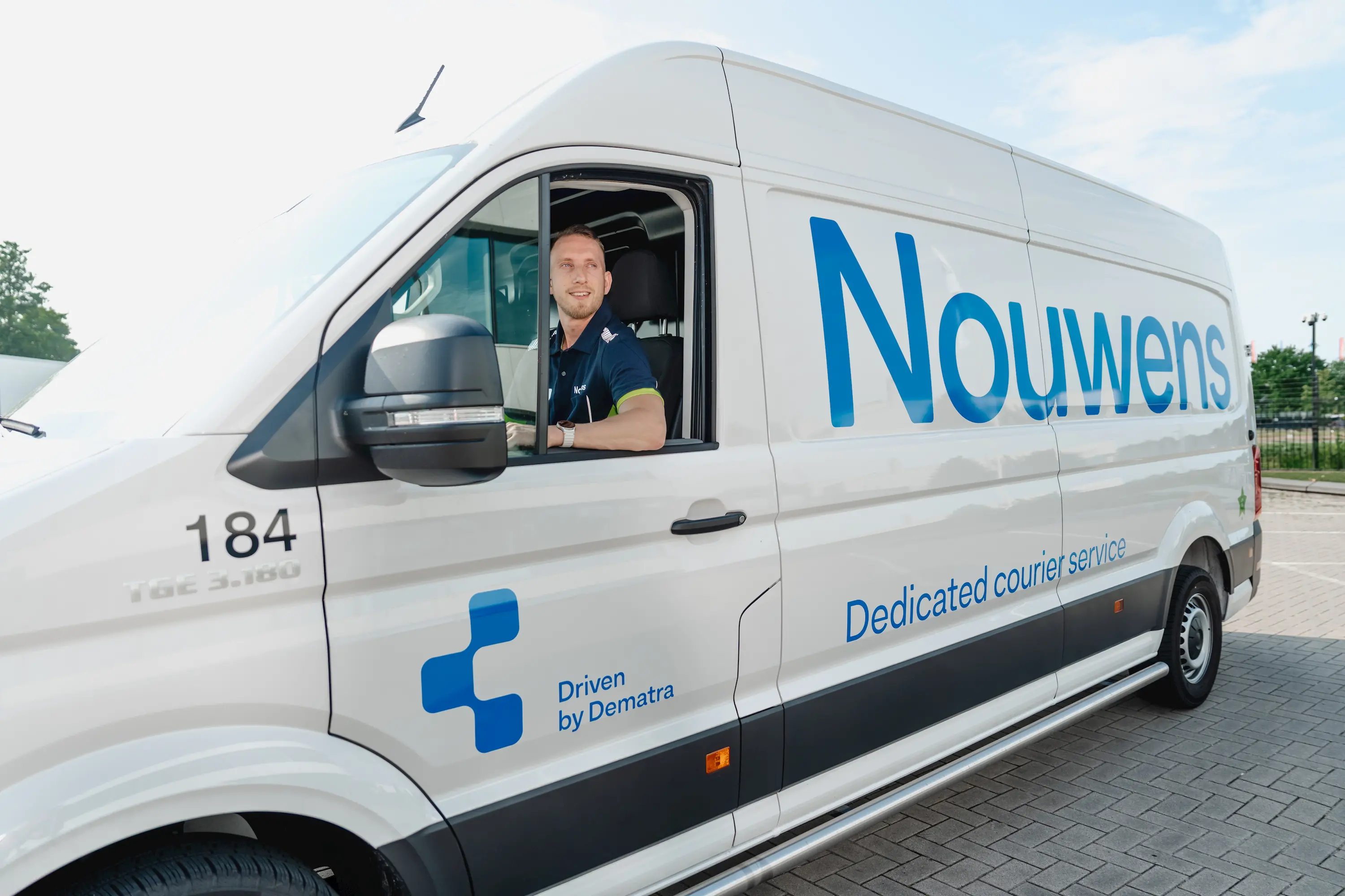 Man in blue shirt sitting in the driver's seat of a white van with 'Nouwens Dedicated courier service' and 'Driven by Dematra' logos.