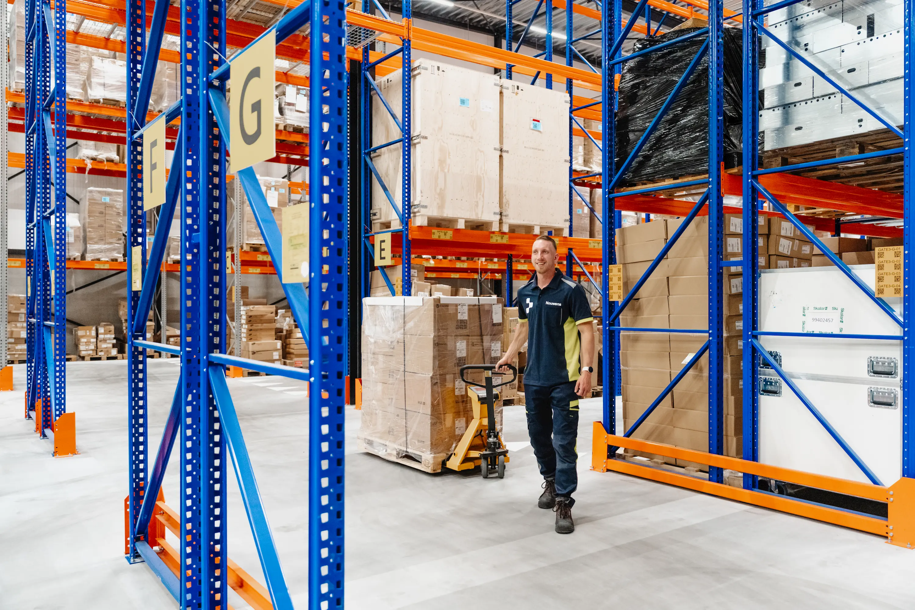 Warehouse worker pushing a pallet jack loaded with boxes through aisles of blue and orange shelving.