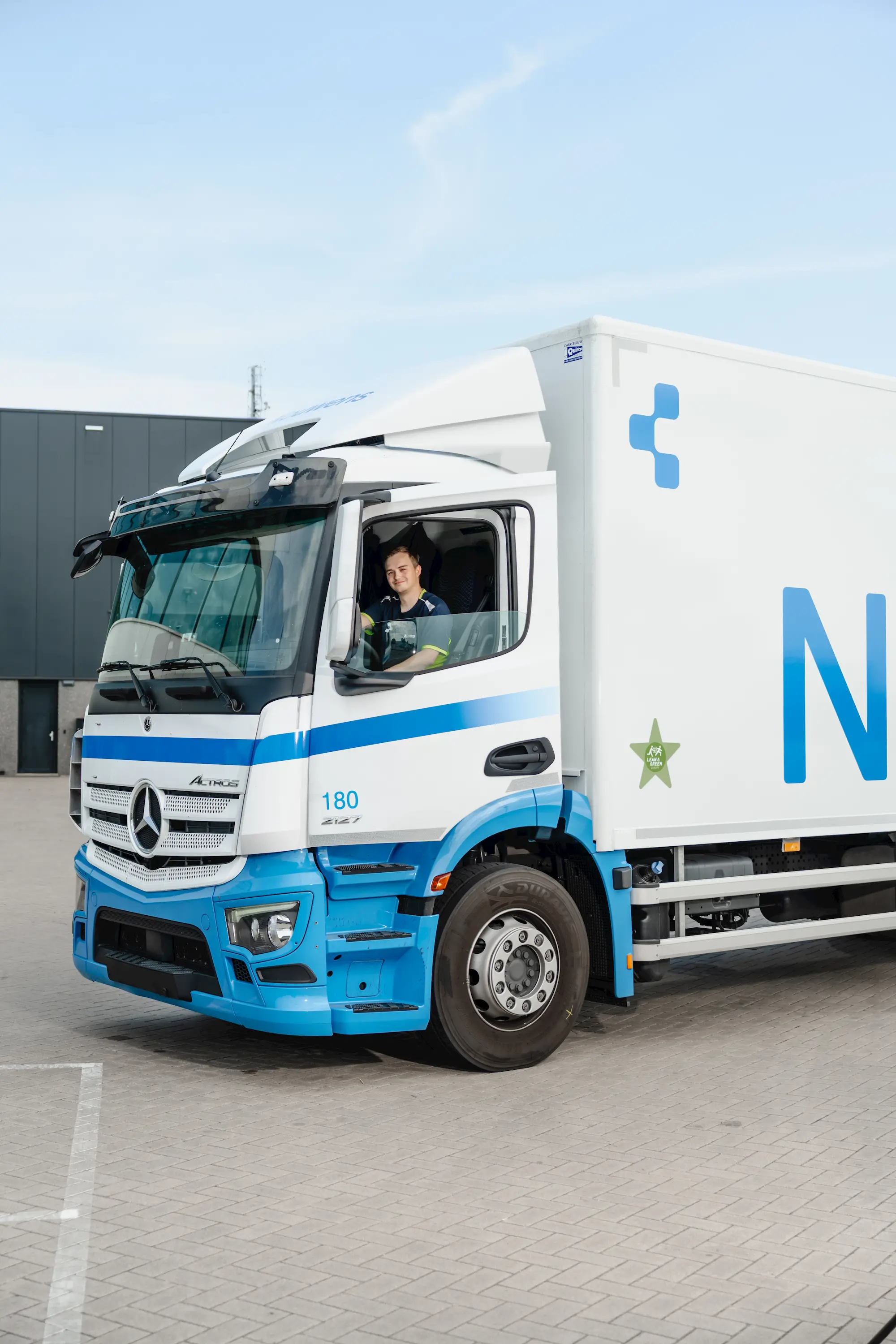 Man smiling and sitting inside a blue and white Mercedes-Benz Actros truck parked on a paved surface.