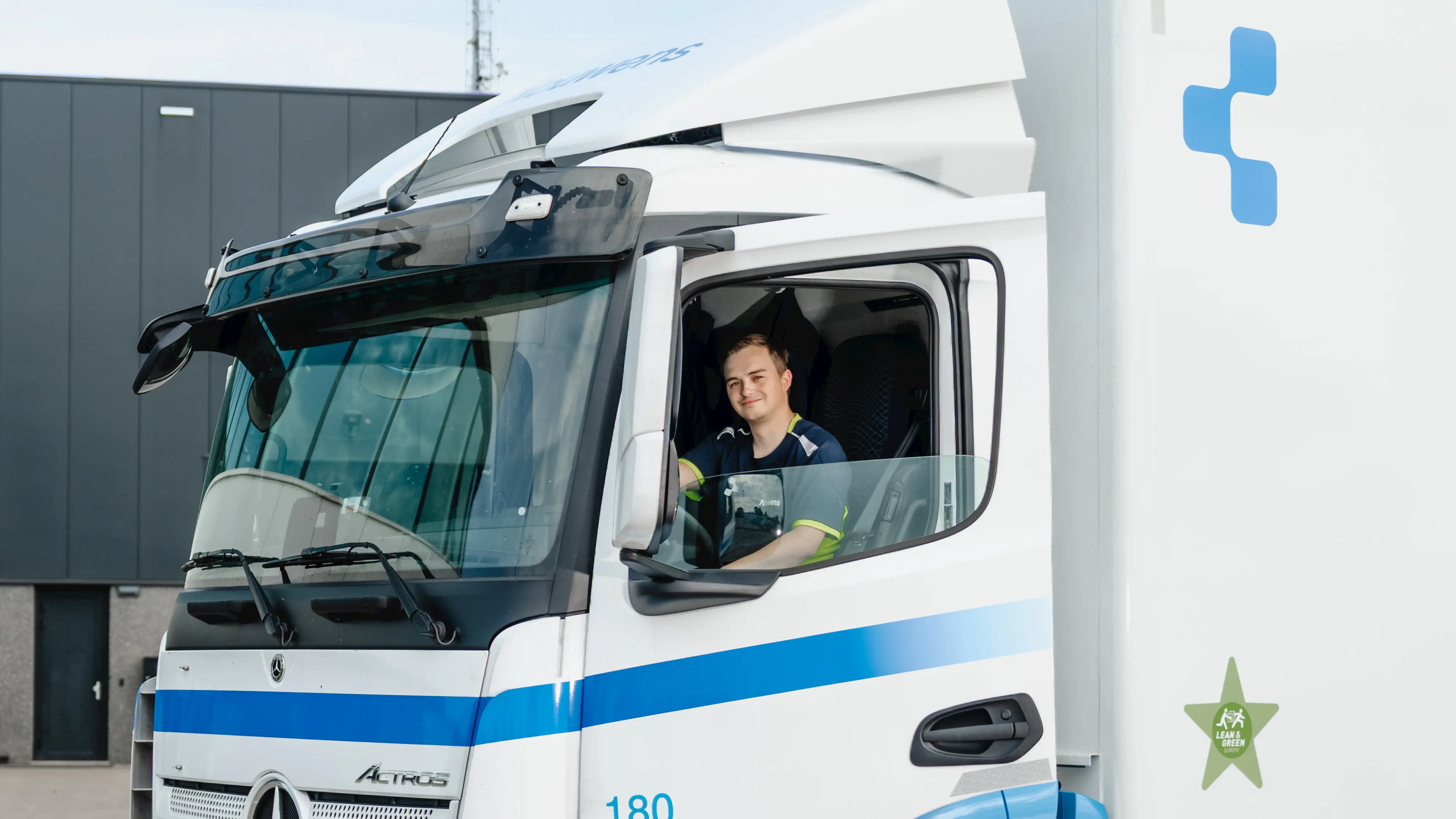 Smiling man sitting in the driver's seat of a white Mercedes Actros truck with blue stripes.