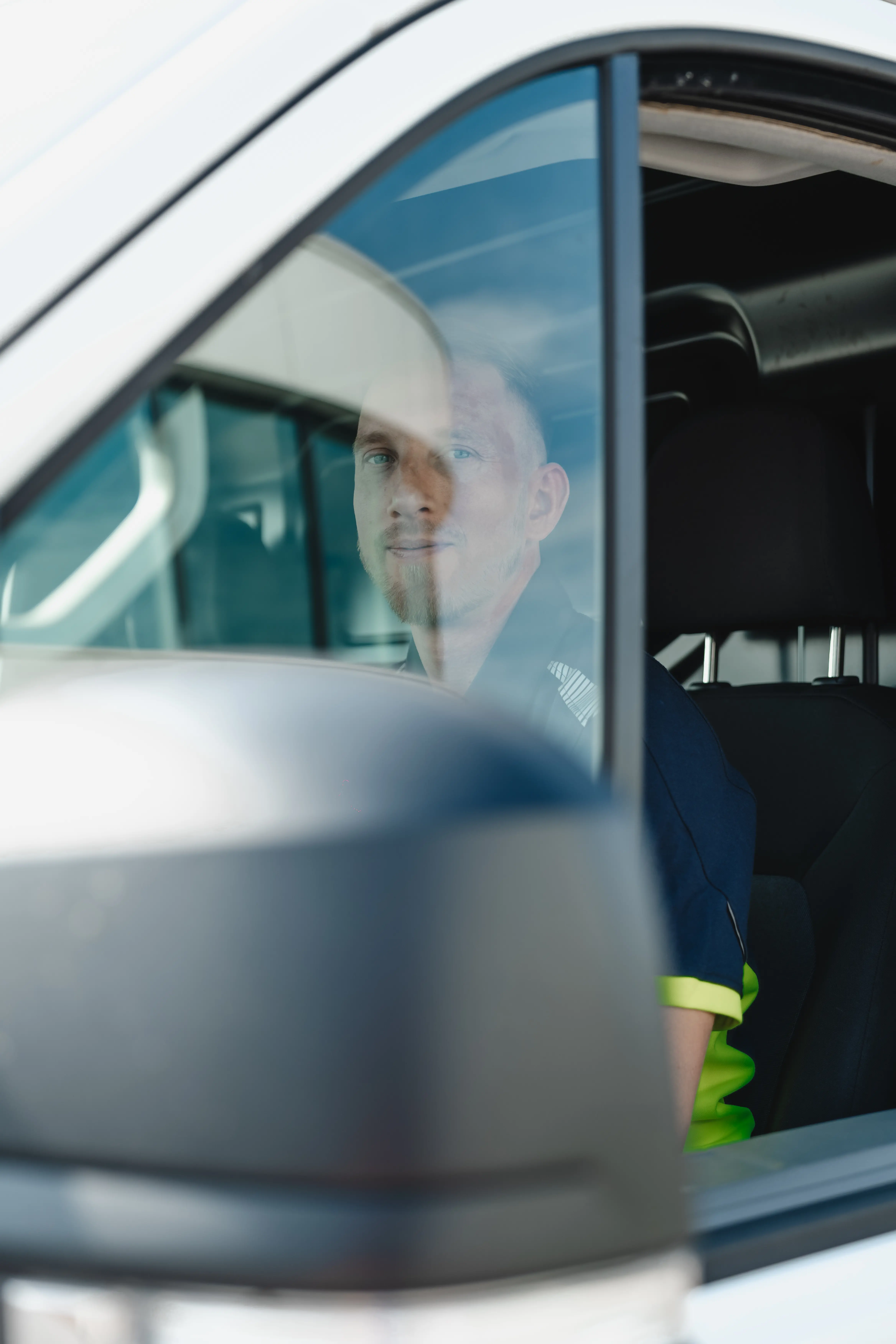Man with light hair and beard sitting in the driver's seat of a vehicle, visible through the window and side mirror.