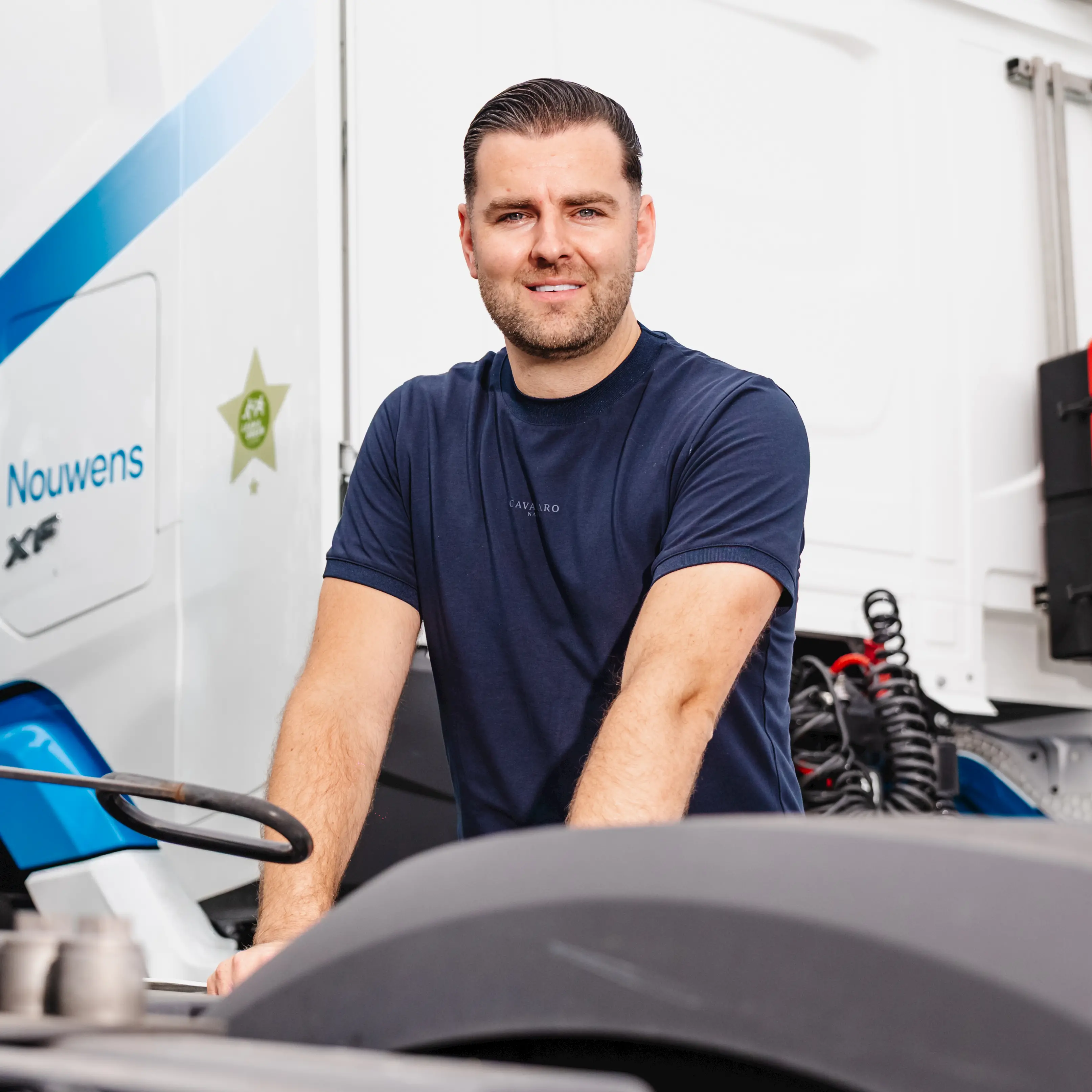 Man in a navy blue t-shirt leaning on a vehicle with a white truck in the background.