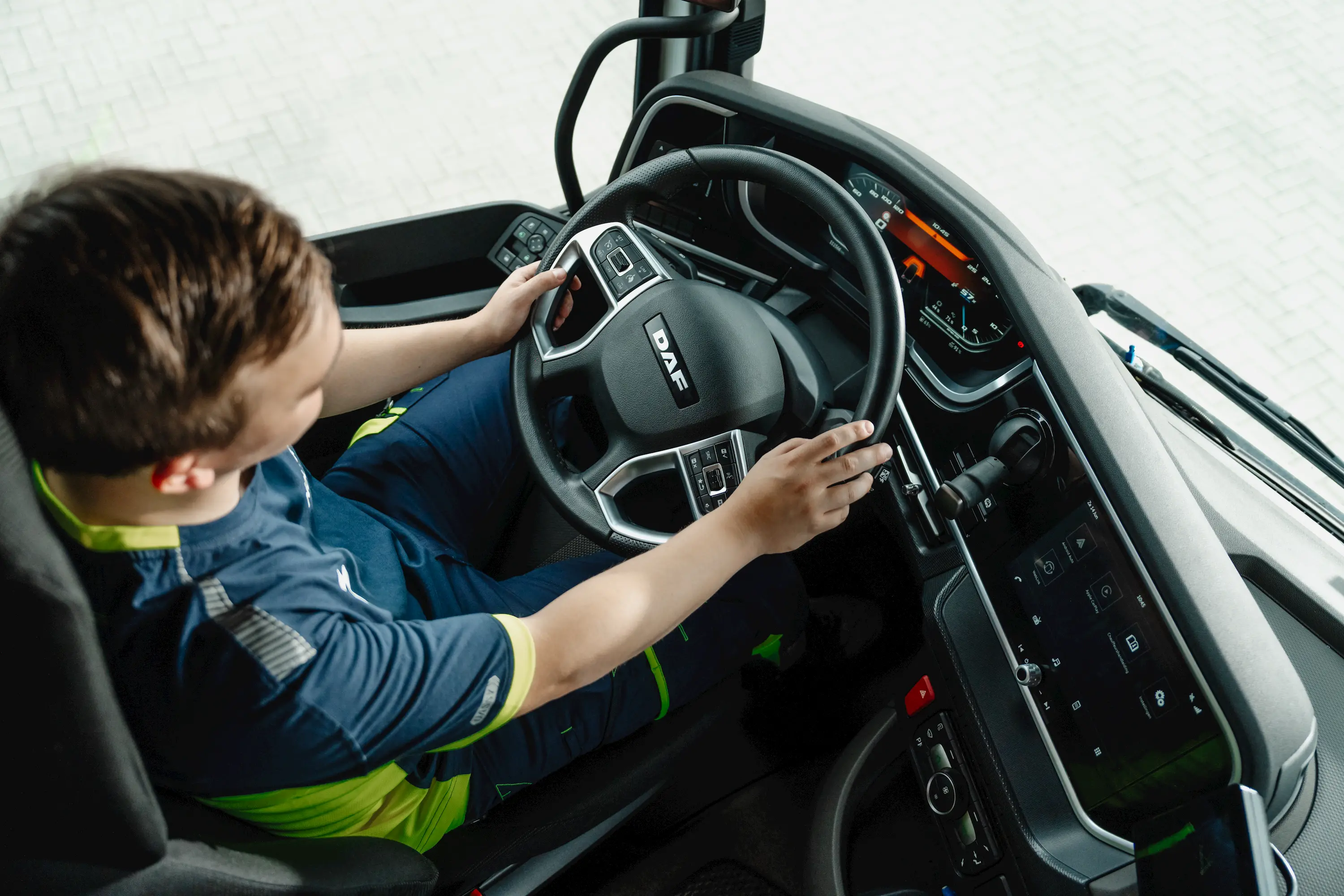 Person seated in a DAF truck cabin holding the steering wheel with dashboard and digital display visible.