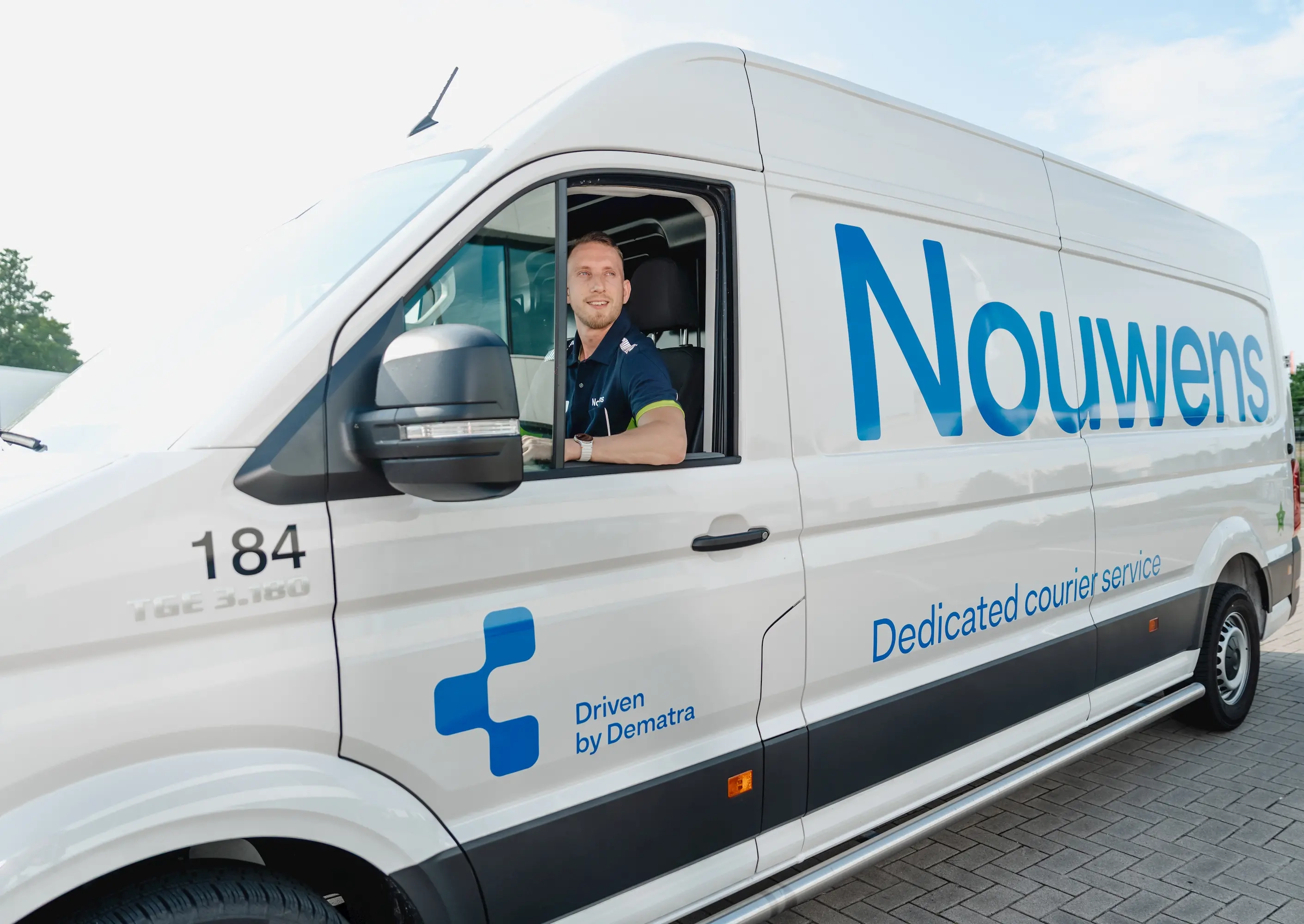 Man wearing a navy uniform sitting inside a white Nouwens courier service van, smiling and looking out the open window.