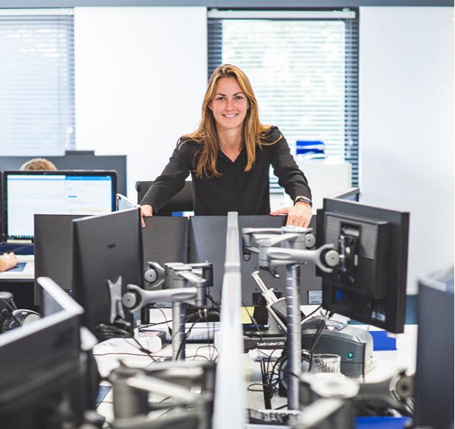 Smiling woman leaning on a desk divider in a modern office with multiple computer monitors.