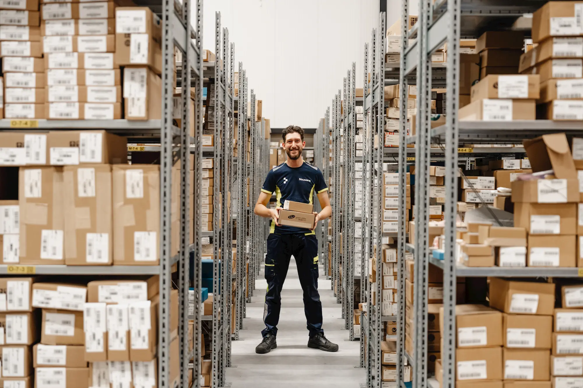 Smiling warehouse worker in uniform holding two cardboard boxes standing between tall shelves filled with packages.