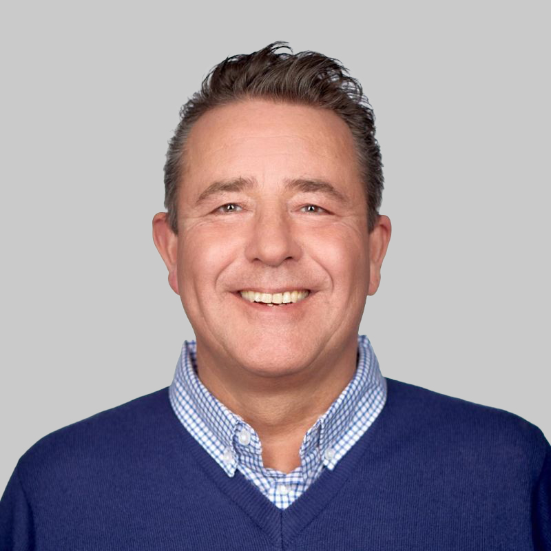 Smiling middle-aged man with short brown hair wearing a blue sweater over a blue and white checkered shirt against a gray background.