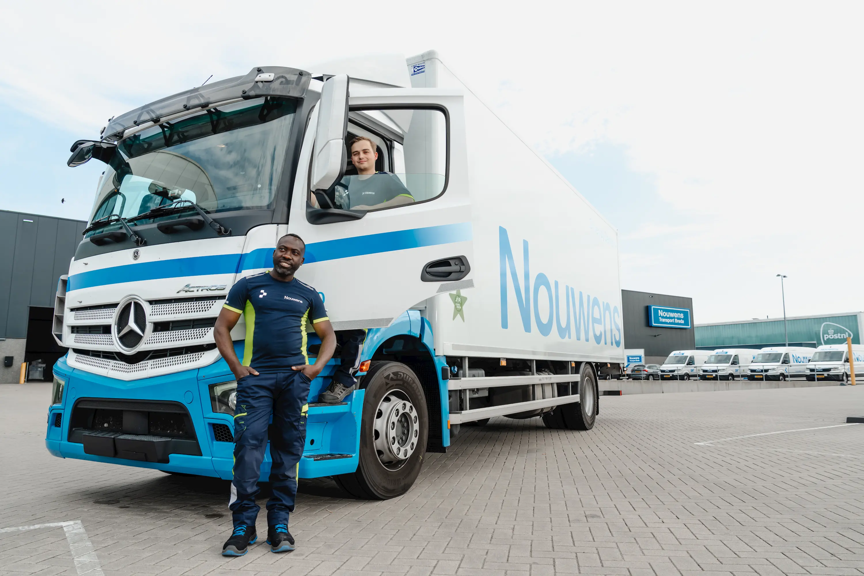 Two delivery workers in uniforms with a white and blue Nouwens delivery truck in an industrial parking area.