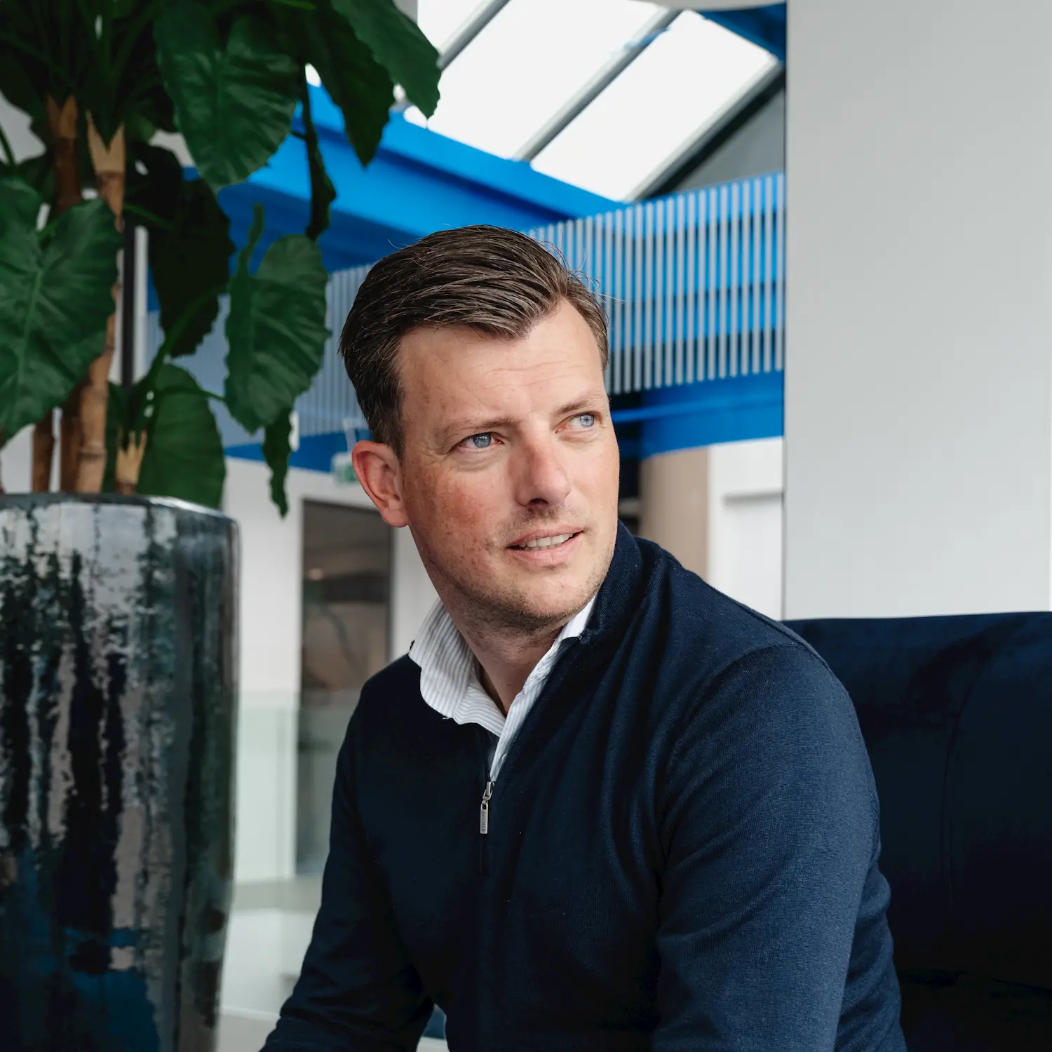 Man with short brown hair wearing a navy blue zip-up sweater over a white collared shirt, sitting indoors near a large plant and modern blue and white interior.