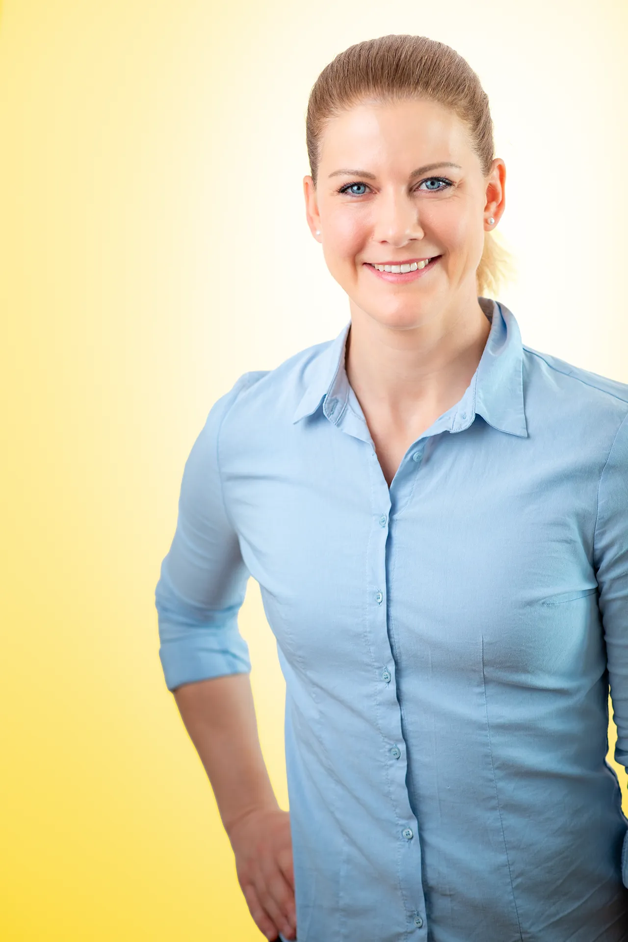 Smiling woman with blue eyes and light brown hair tied back, wearing a light blue button-down shirt, standing against a yellow gradient background.
