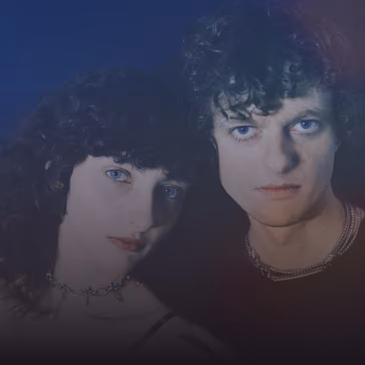 Un homme et une femme aux yeux bleus, aux cheveux bouclés, posant près l'un de l'autre sur fond sombre.