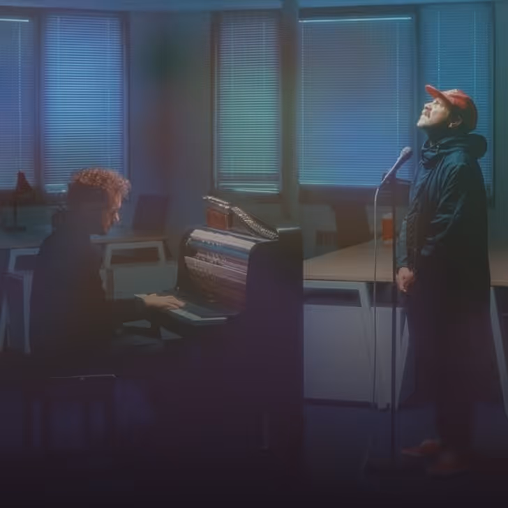 Un homme joue du piano tandis qu'un autre chante debout près d'un microphone dans une pièce sombre.