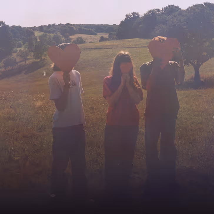 Trois personnes tiennent des cœurs rouges devant leur visage dans un champ sous un ciel clair.