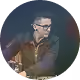Homme portant des lunettes et une veste, regardant vers la droite dans un fond circulaire sombre.