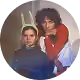 Deux jeunes adultes aux cheveux bouclés, l'un en chemise bleue, l'autre en pull rouge, posent ensemble.