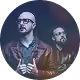 Deux hommes souriants portant des lunettes dans une lumière colorée sombre.