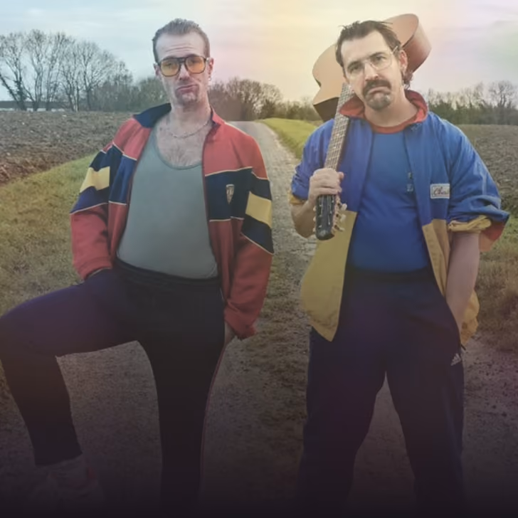 Deux hommes posent sur un chemin rural, l'un tient une guitare sur l'épaule.