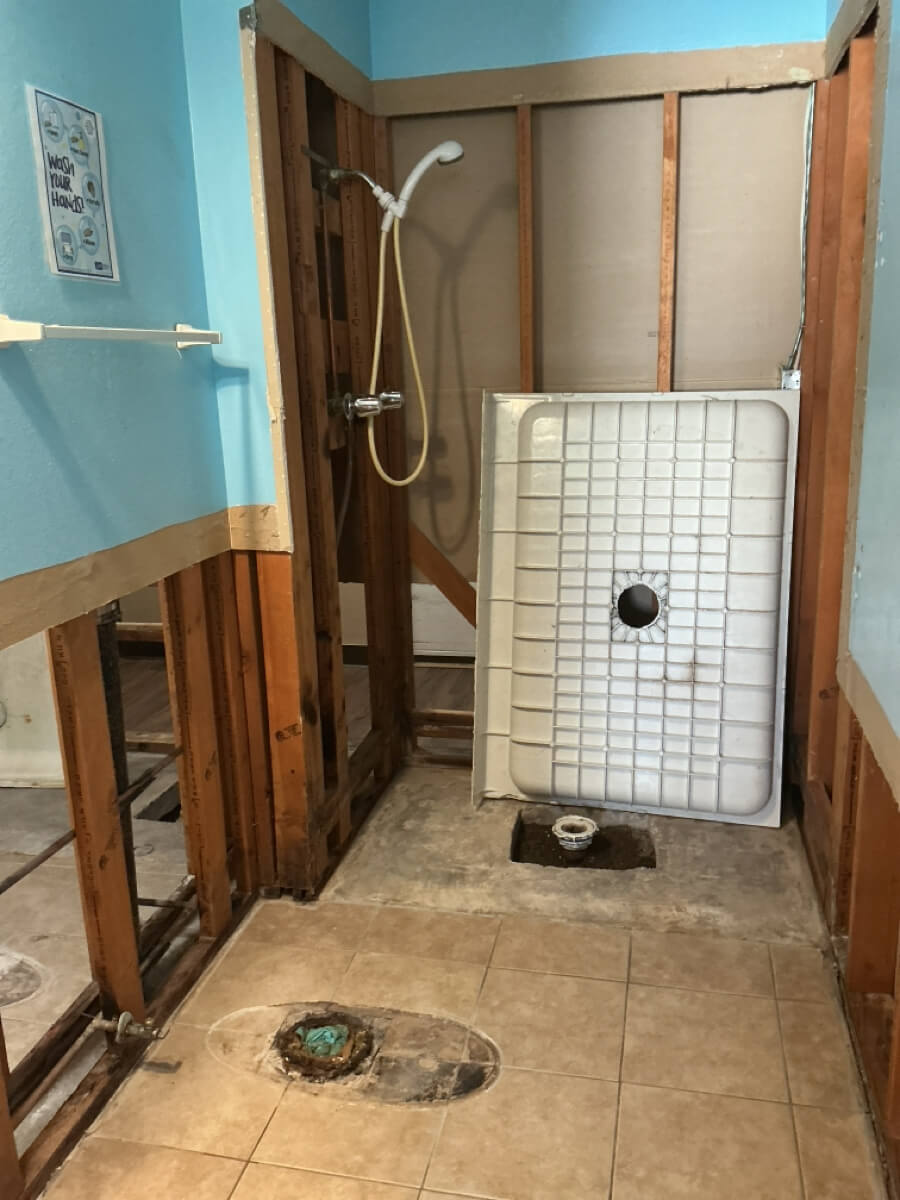 Bathroom under renovation with exposed wooden framing, a dismantled shower area, blue walls, and beige tiles.