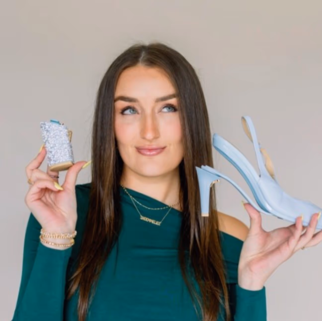Woman in green top holding a light blue high heel shoe in one hand and its sparkling heel in the other, looking thoughtfully upward.