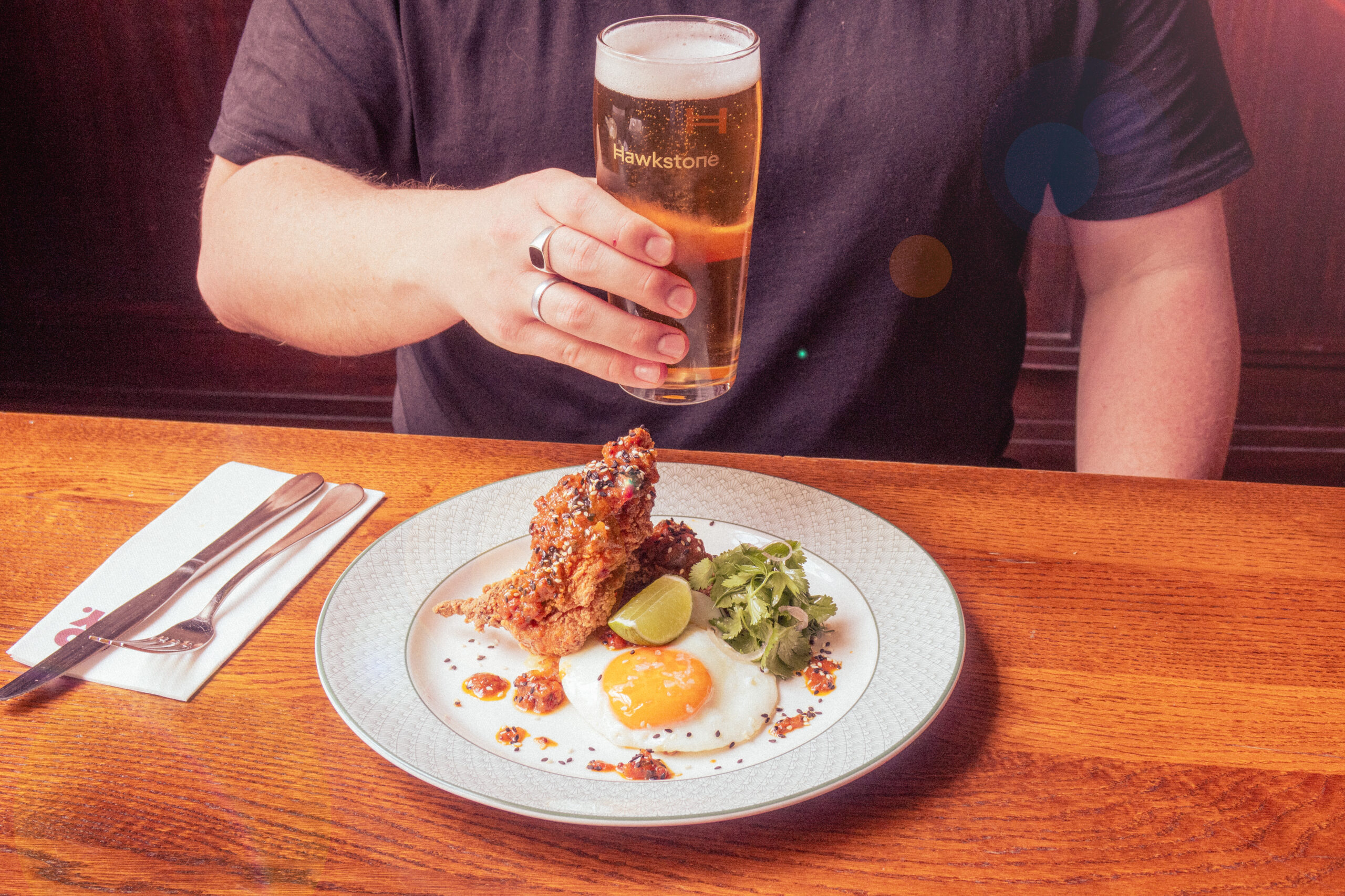 A person holds a pint of beer above a plate with fried chicken, a sunny-side-up egg, lime wedge, greens, and sauce, on a wooden table with utensils and a napkin on the side.