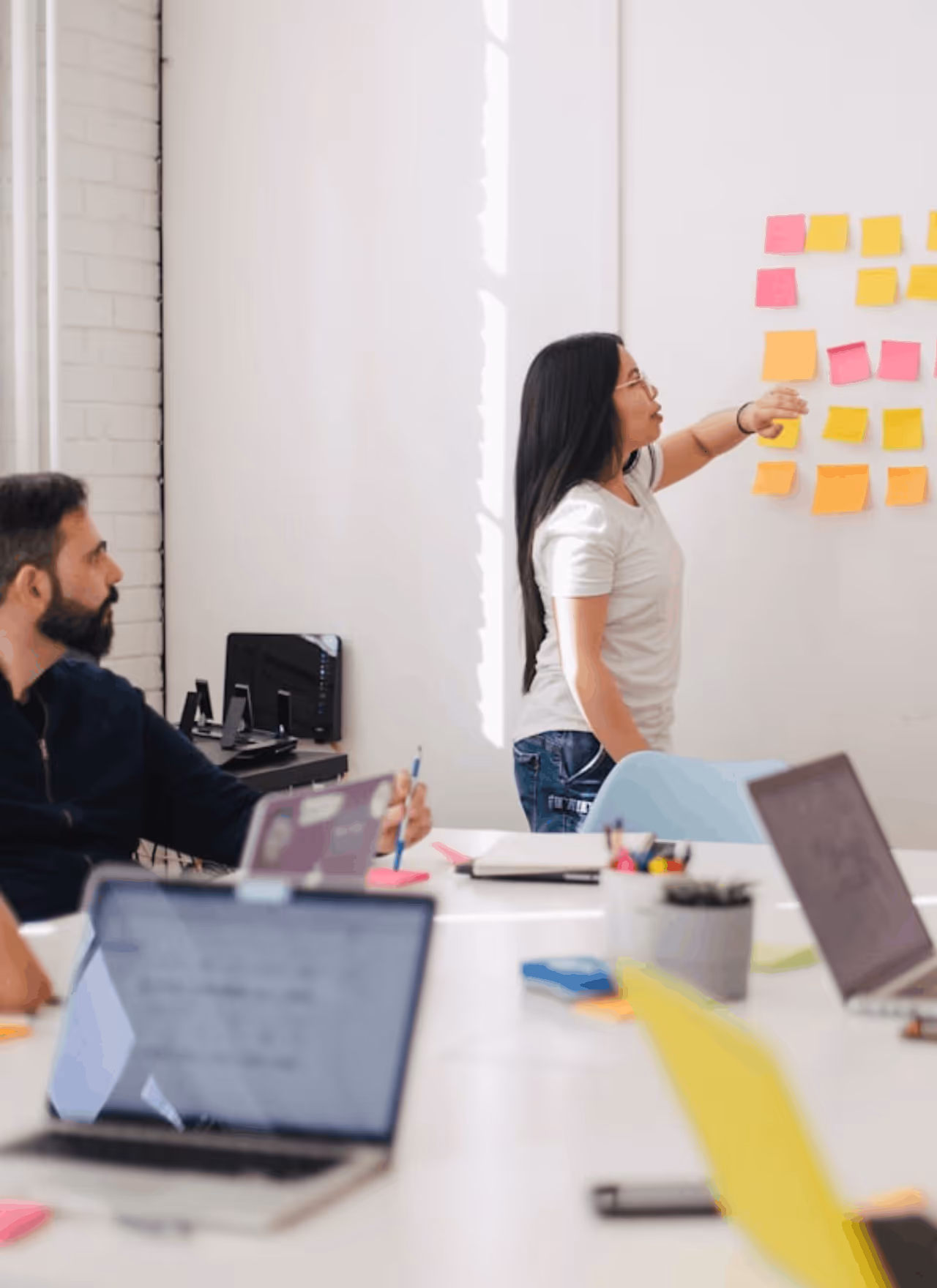 Mujer señalando notas adhesivas de colores en una pared mientras dos personas la escuchan en una reunión de oficina.