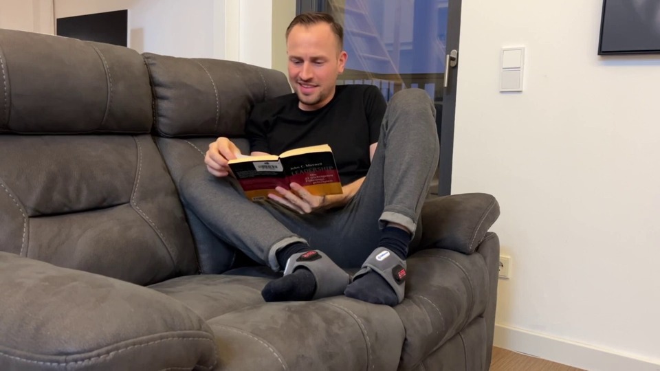 A man reading on a grey sofa while wearing EMSense wraps on both feet.