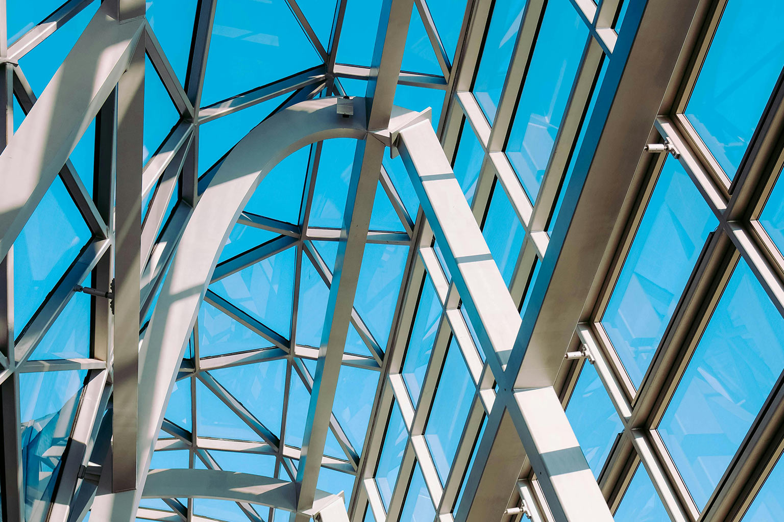 Interior view of a modern industrial glass-and-steel structure with intersecting beams and bright blue skylights, representing innovation and safety-focused design.