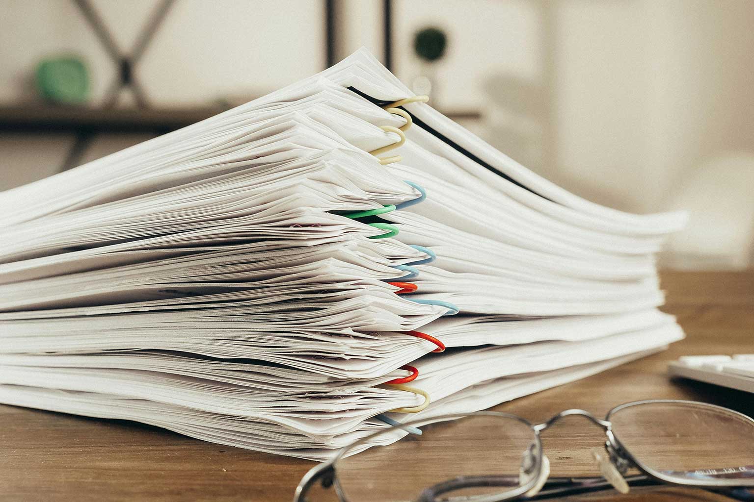 Understanding and Accessing Current Galvanizing Standards — a stack of organized paper documents clipped with colored paperclips on a desk, with reading glasses in the foreground in an office setting.
