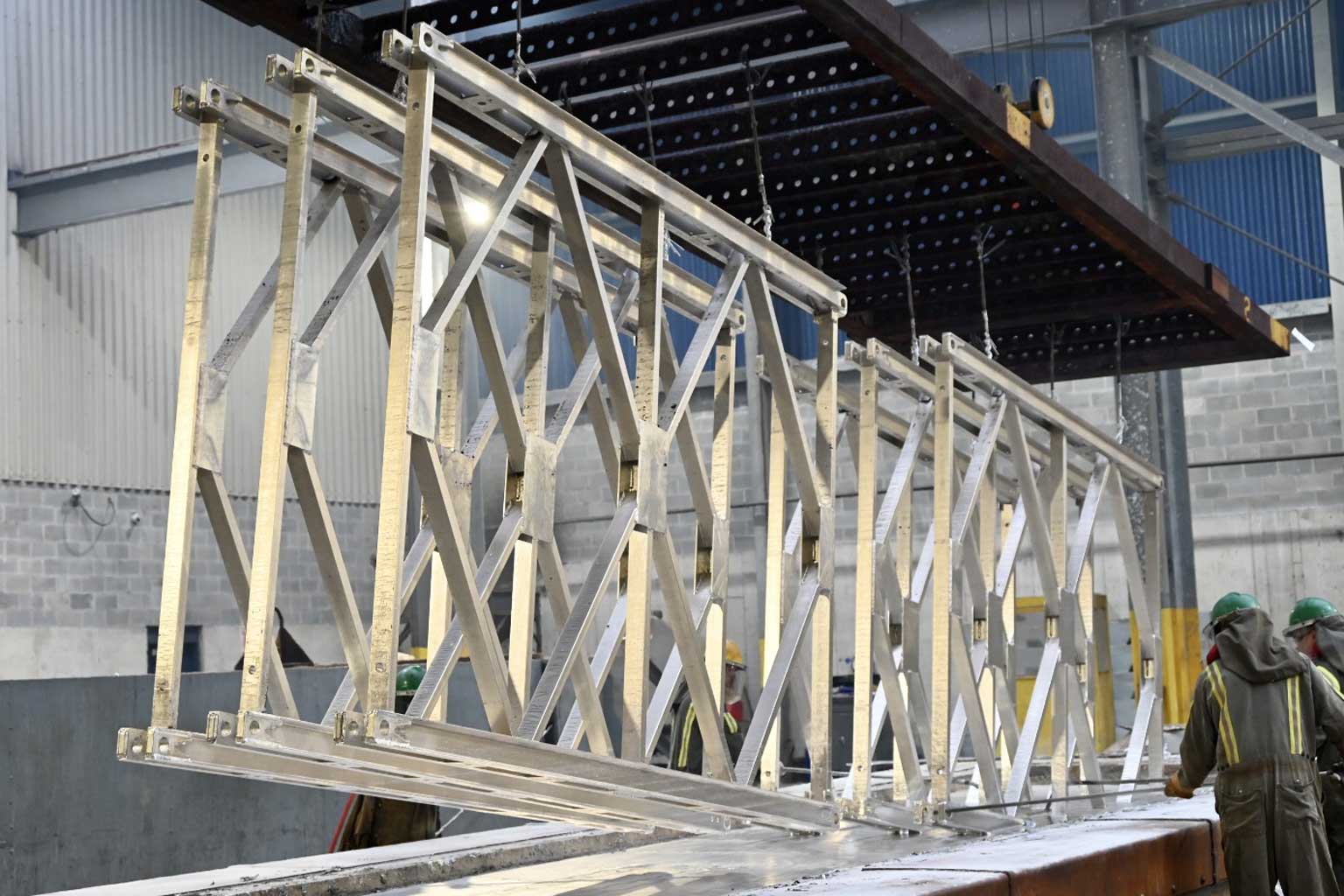 Series of tall lattice-like steel frames being lifted from a large galvanizing kettle inside a plant, illustrating progressive dipping of long articles.