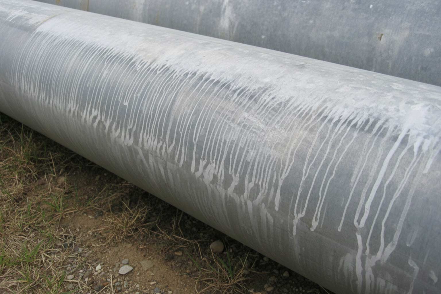 Large galvanized steel pipe lying outdoors on the ground with streaked white runoff marks along its surface, demonstrating coating and cooling effects.