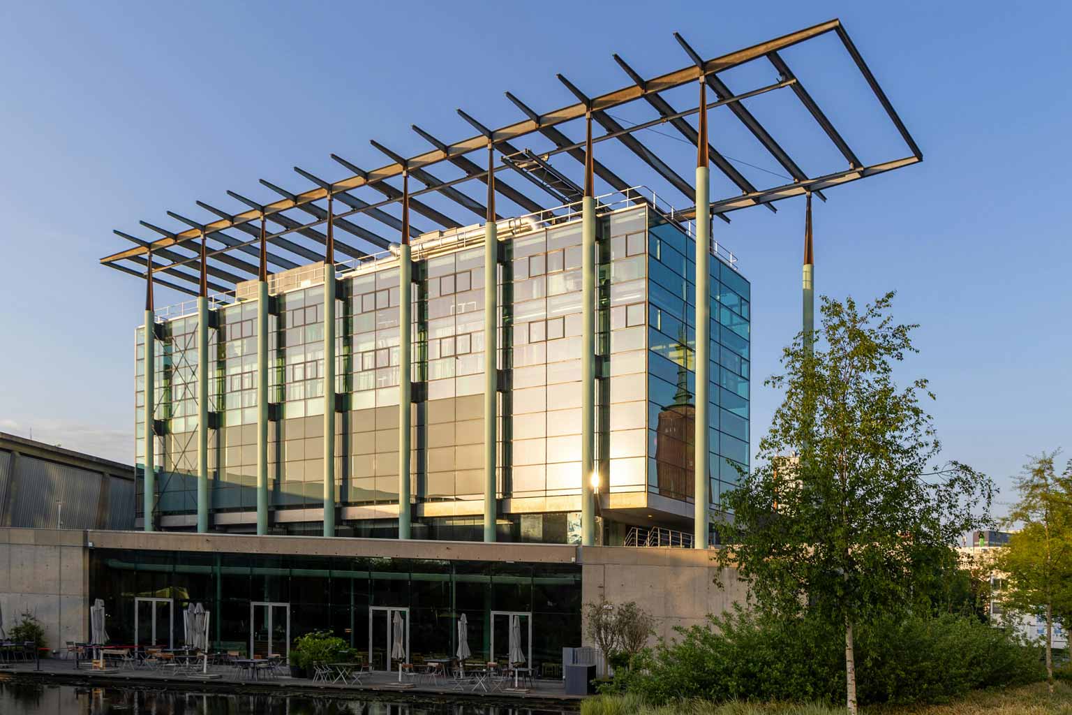 Modern glass office building with exposed galvanized steel columns and roof framing, reflecting evening light above a ground-level terrace and landscaping.