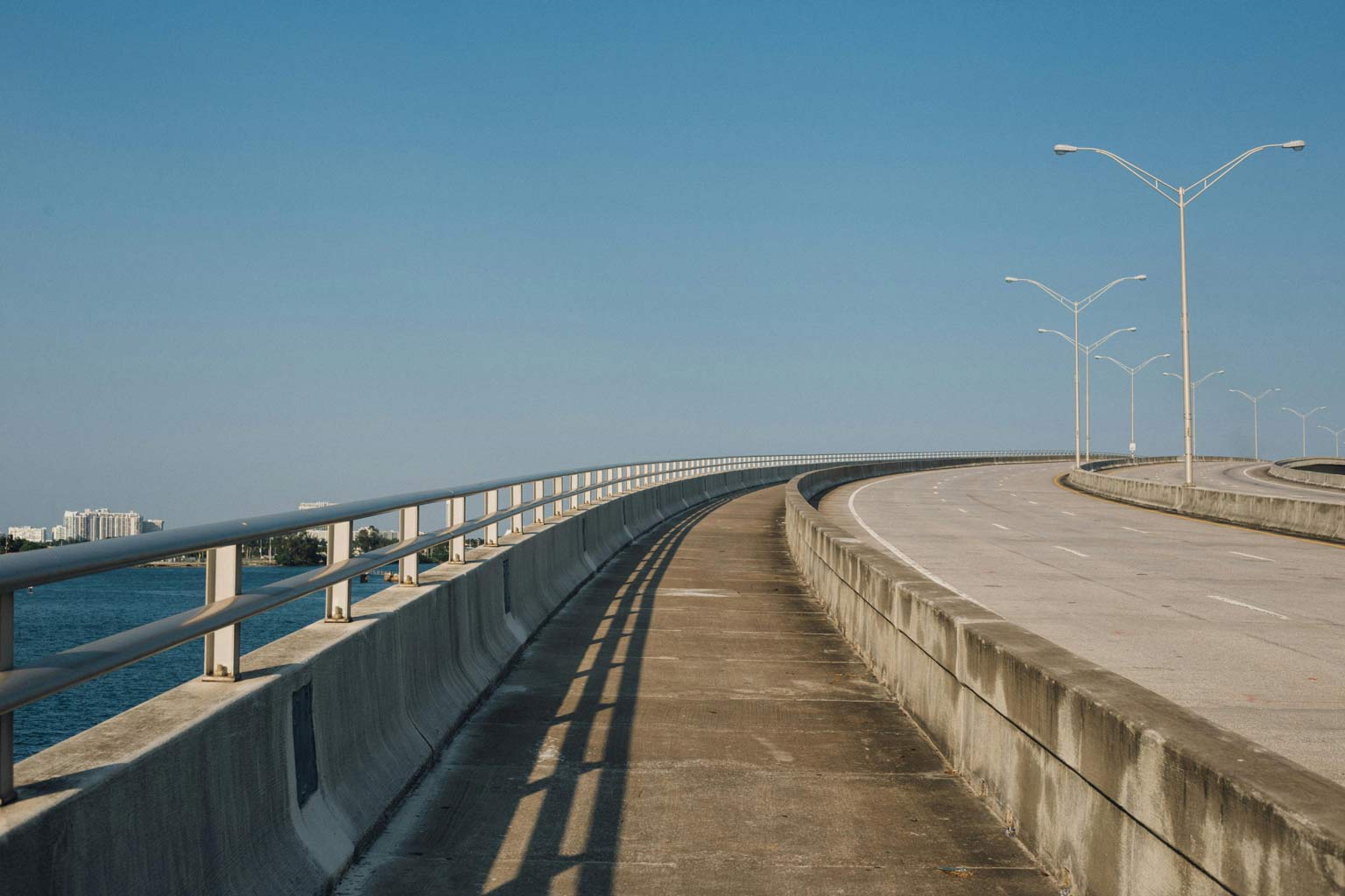 Curving concrete bridge and roadway with metal guardrails and light poles, stretching over water under a clear blue sky.