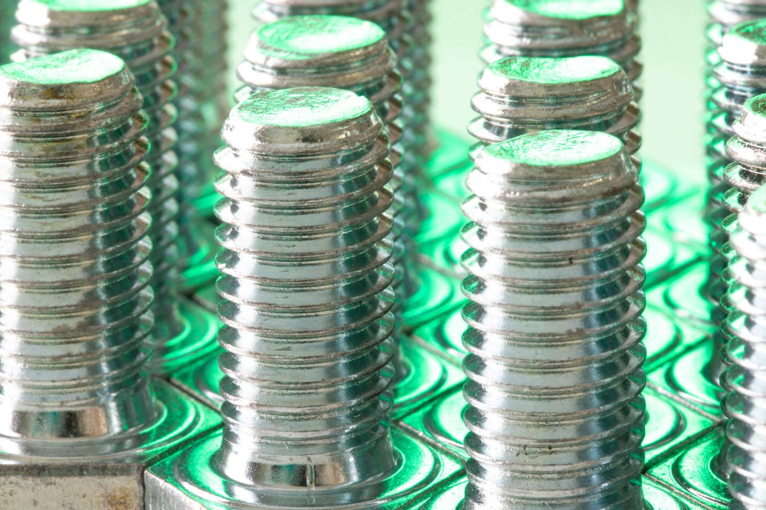 Rows of shiny threaded bolts standing upright, reflecting green light that highlights the metallic finish and uniform threads.
