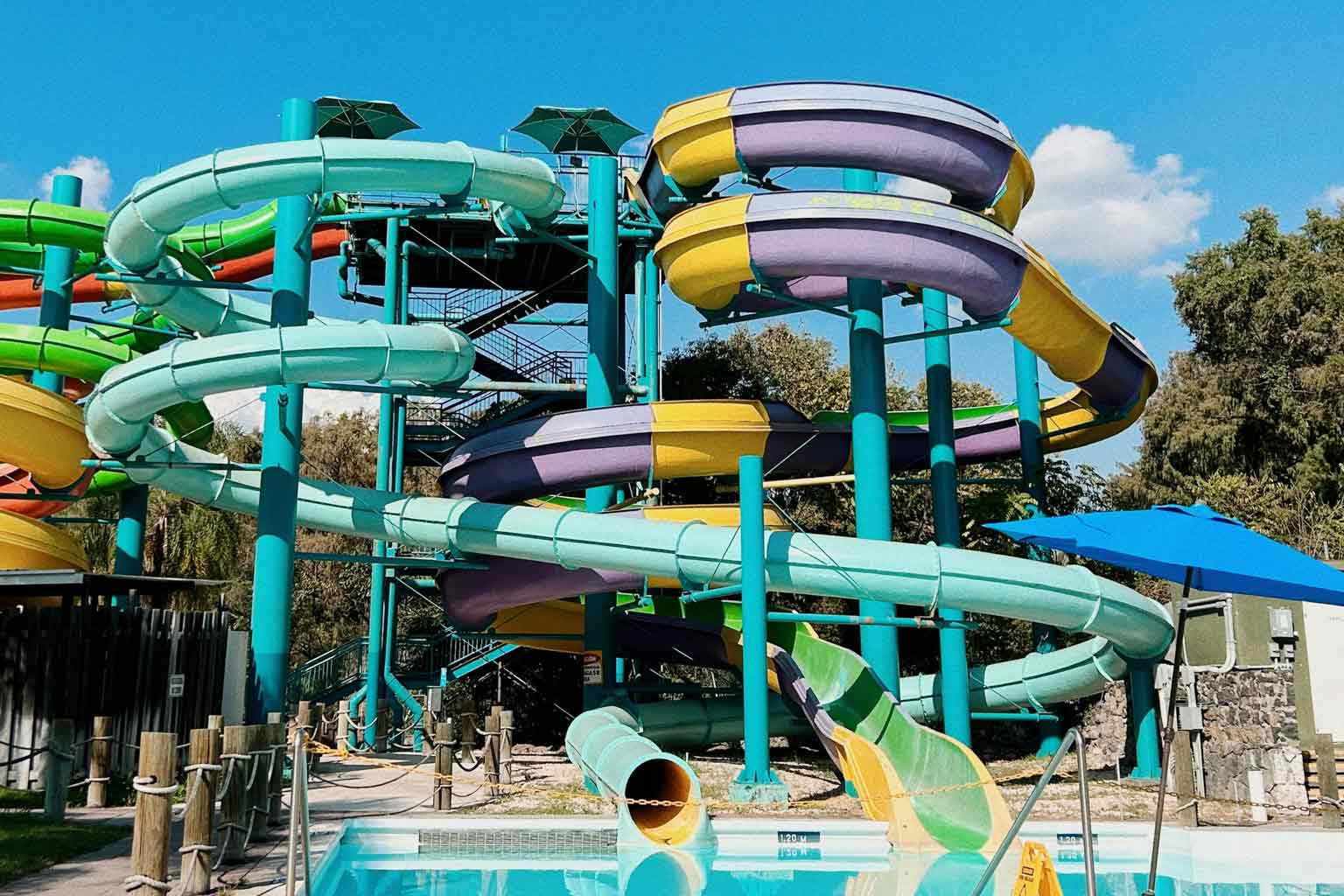 Colorful water park with looping enclosed water slides supported by teal steel columns above a bright blue swimming pool.