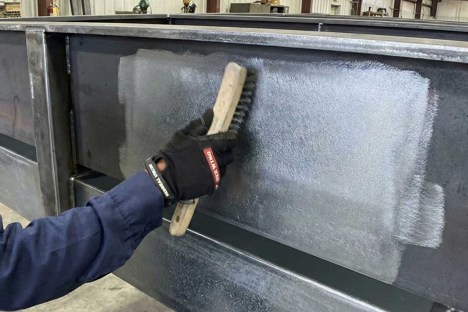 Worker using a wire brush to clean or prepare the galvanized steel surface of a large beam inside a fabrication facility.
