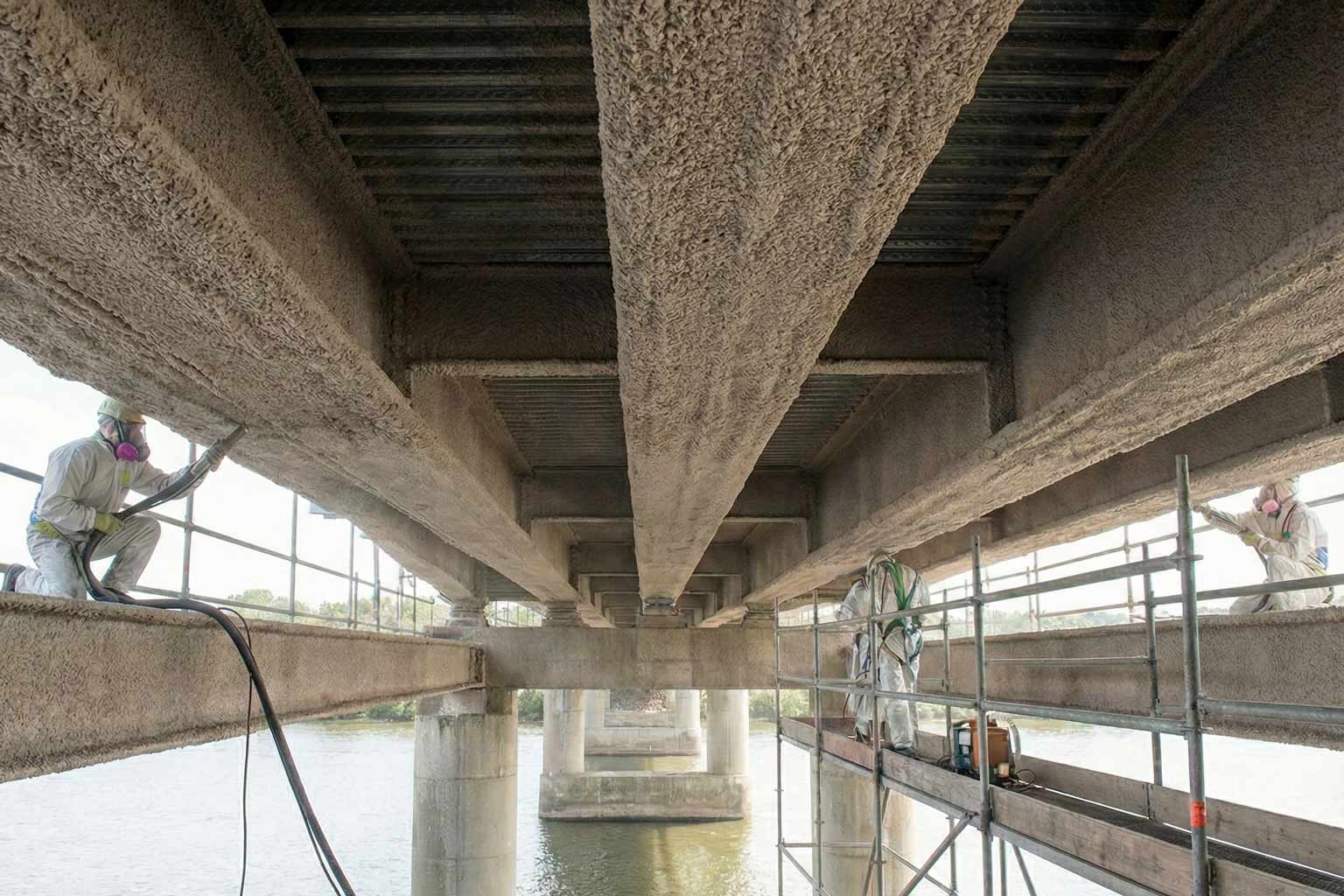 Workers in protective suits applying fireproofing material under a steel bridge structure, demonstrating surface preparation needs before passive fireproofing.