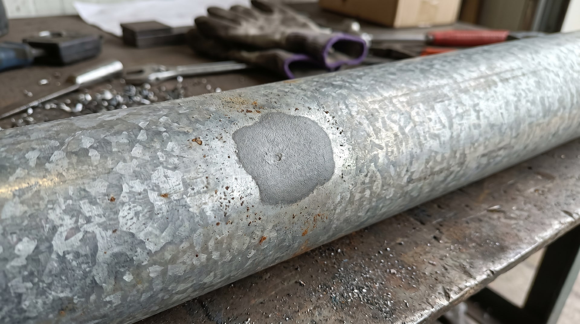 Close-up of a galvanized steel pipe section showing a matte gray flux inclusion spot where the zinc coating failed to form, under industrial workshop lighting.