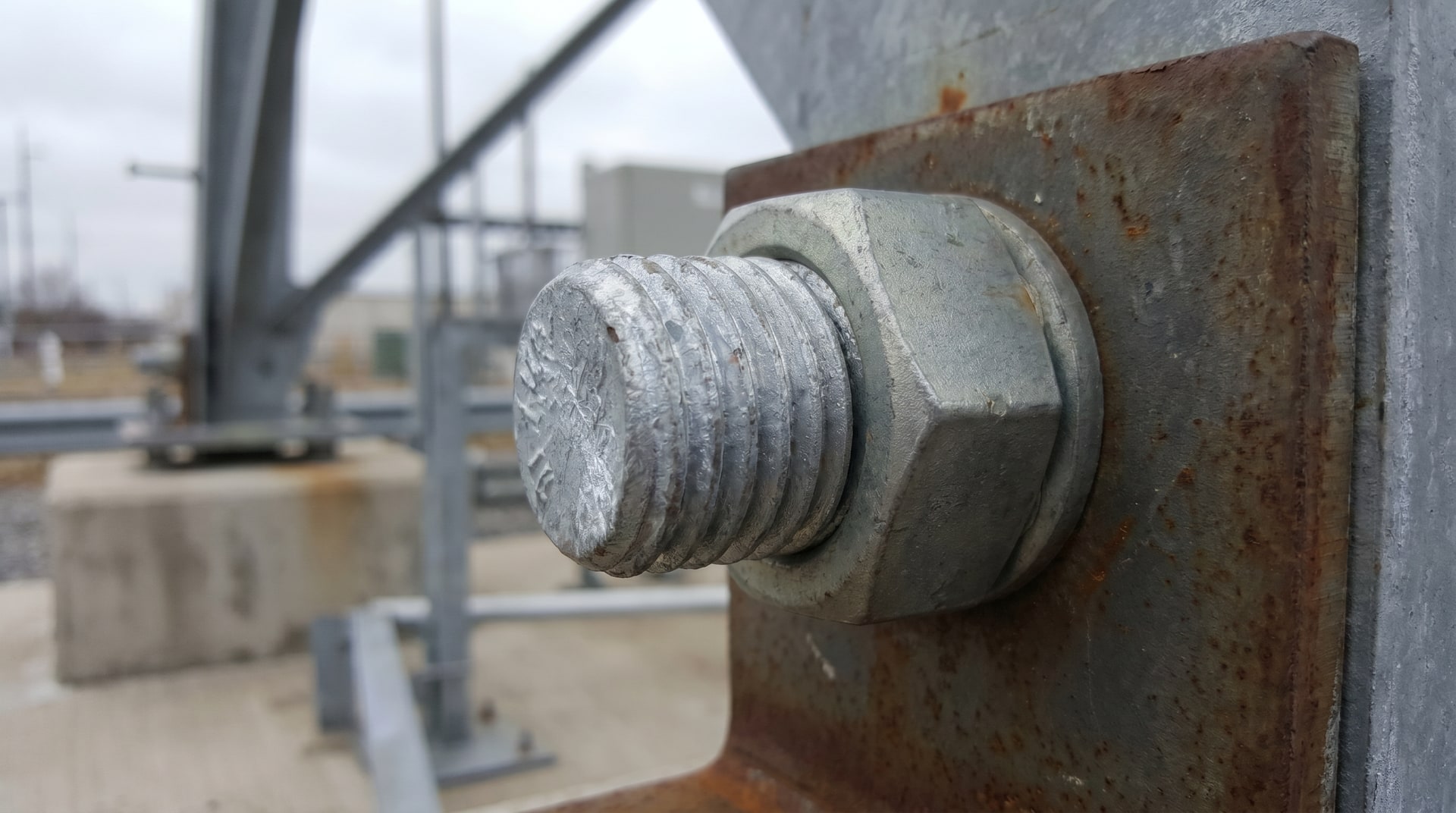 Close-up of a hot-dip galvanized structural bolt and mechanically galvanized nut assembly on a steel connection plate, showing the bright zinc-coated surfaces in an outdoor industrial setting.