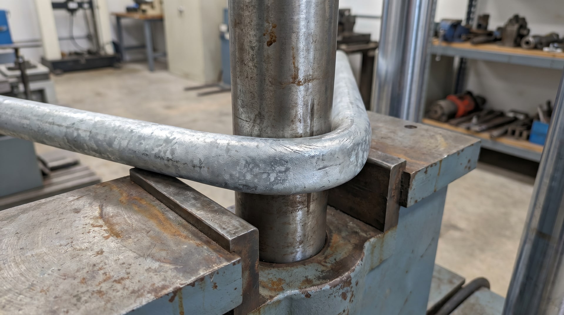 Metallurgist performing a bend ductility test on a hot-dip galvanized steel bar in a mechanical testing laboratory, with zinc coating visible on the specimen surface.