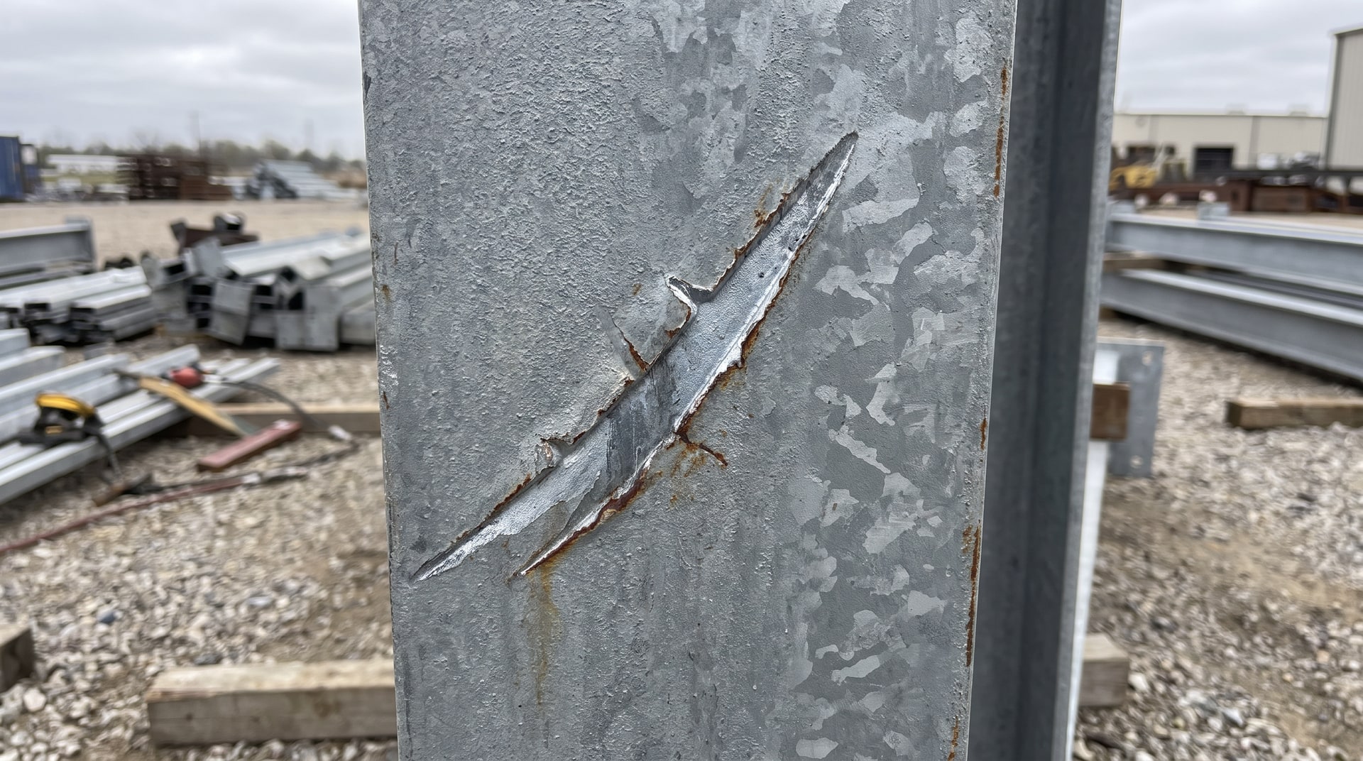 Close-up of a scratch mark in a hot-dip galvanized steel surface showing exposed bare steel beneath the zinc coating, photographed under natural daylight on a fabrication site.