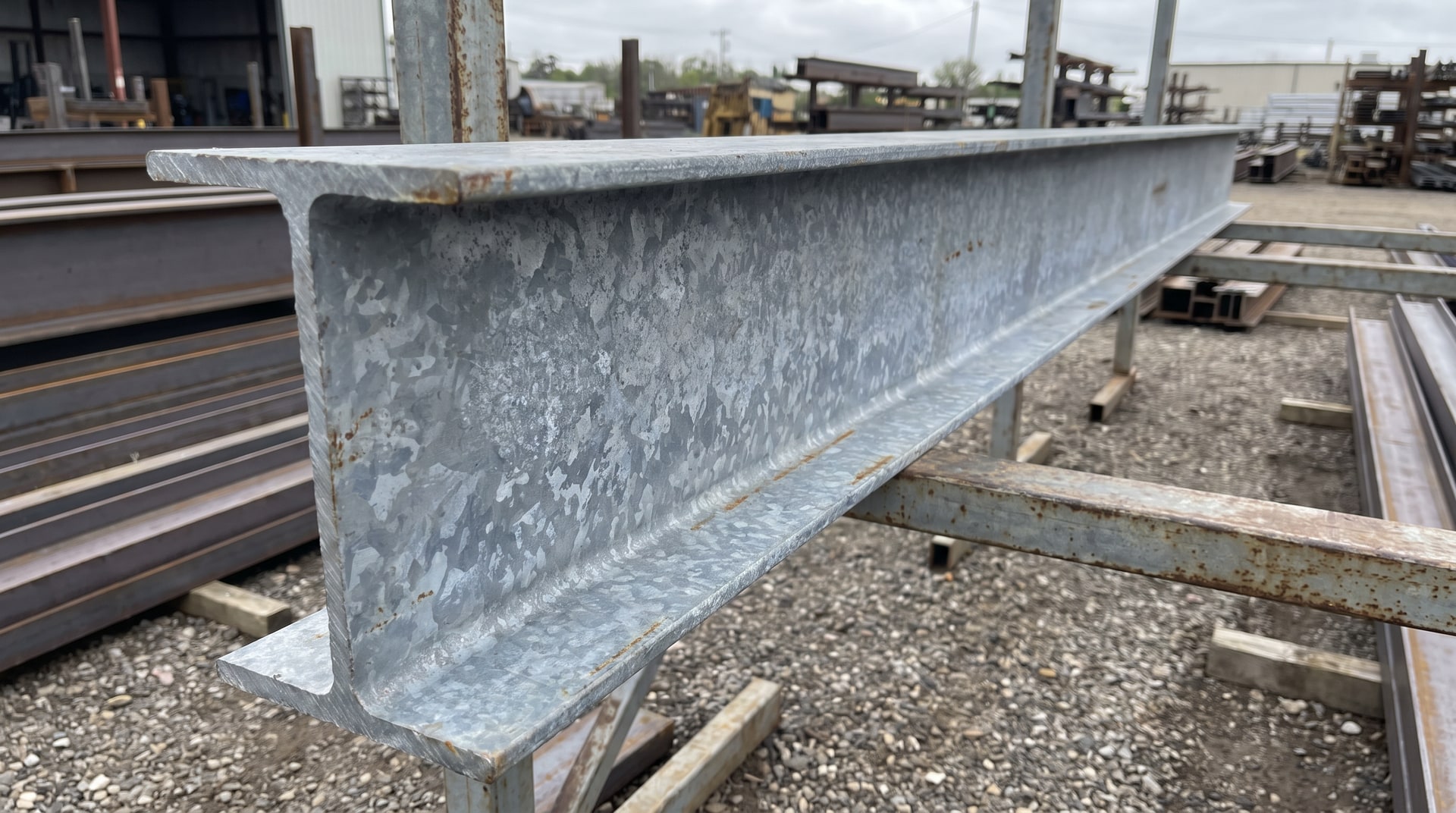 Close-up of a hot-dip galvanized structural steel beam with a visible zinc coating surface, resting on a fabrication yard rack under natural daylight.