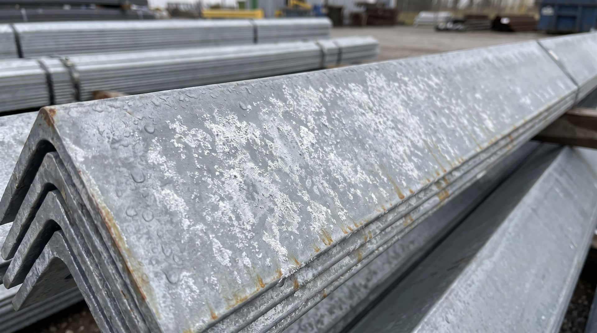 Close-up of galvanized steel angle iron showing white powdery wet storage stain corrosion product on the zinc coating surface, stored outdoors in a humid environment.