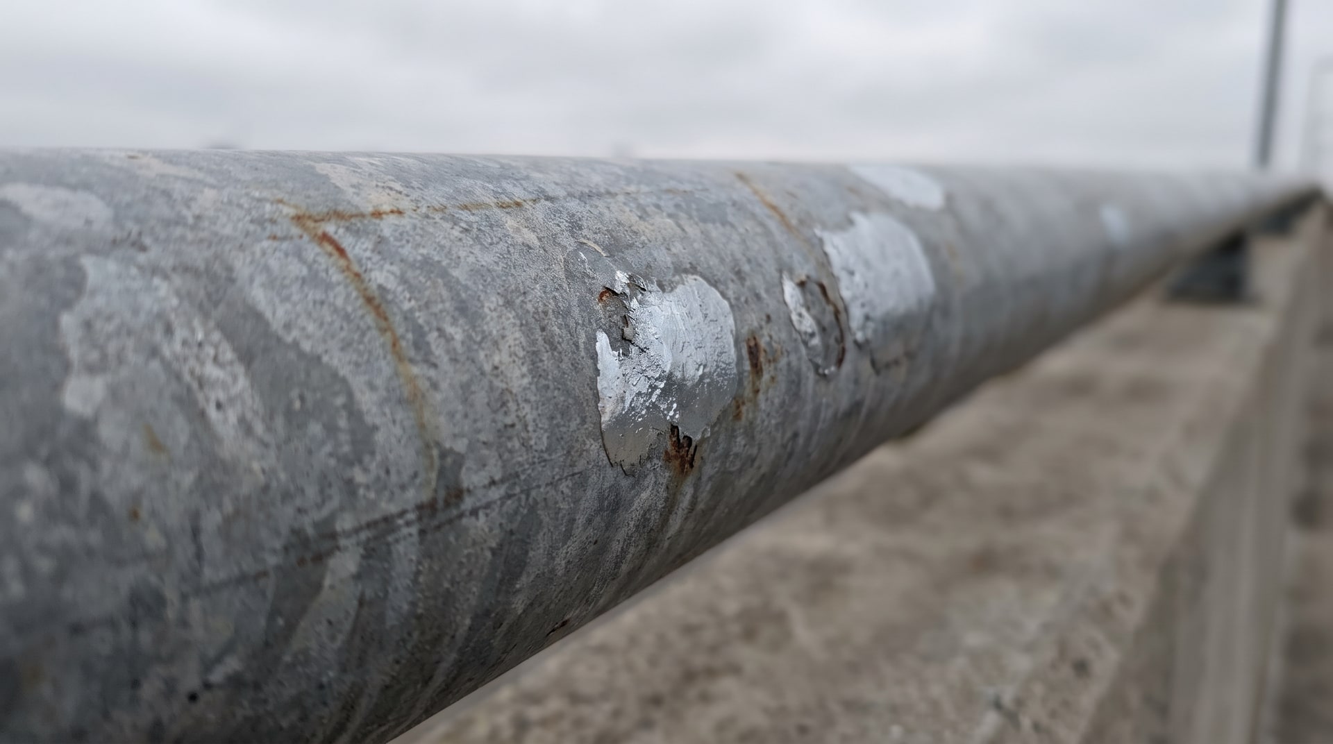 Close-up of a weathered hot-dip galvanized pedestrian handrail on a bridge, showing matte gray patina alongside visibly shinier zinc-rich paint repair patches under overcast daylight.