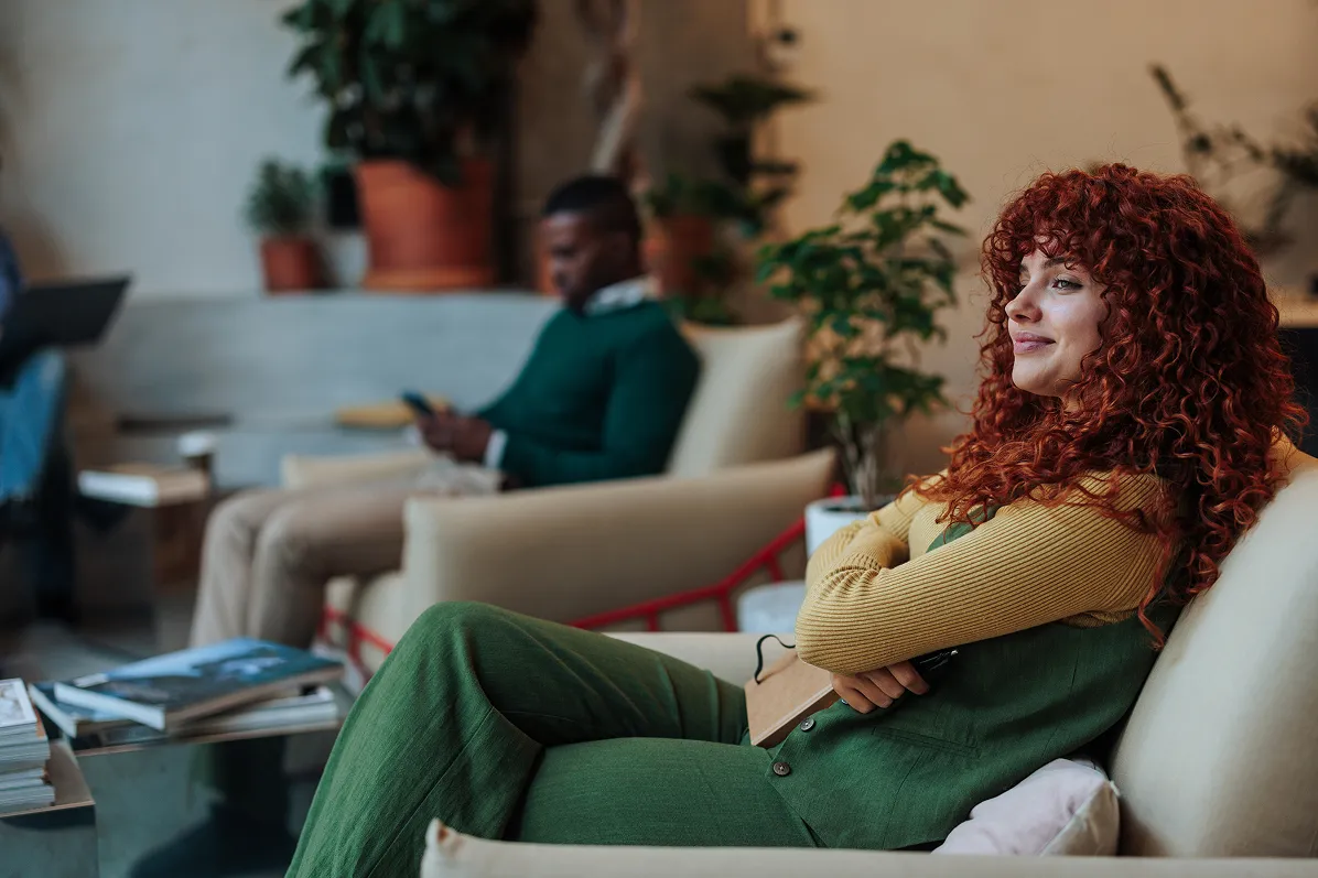 A woman with curly red hair sits on a couch, smiling and holding a notebook.