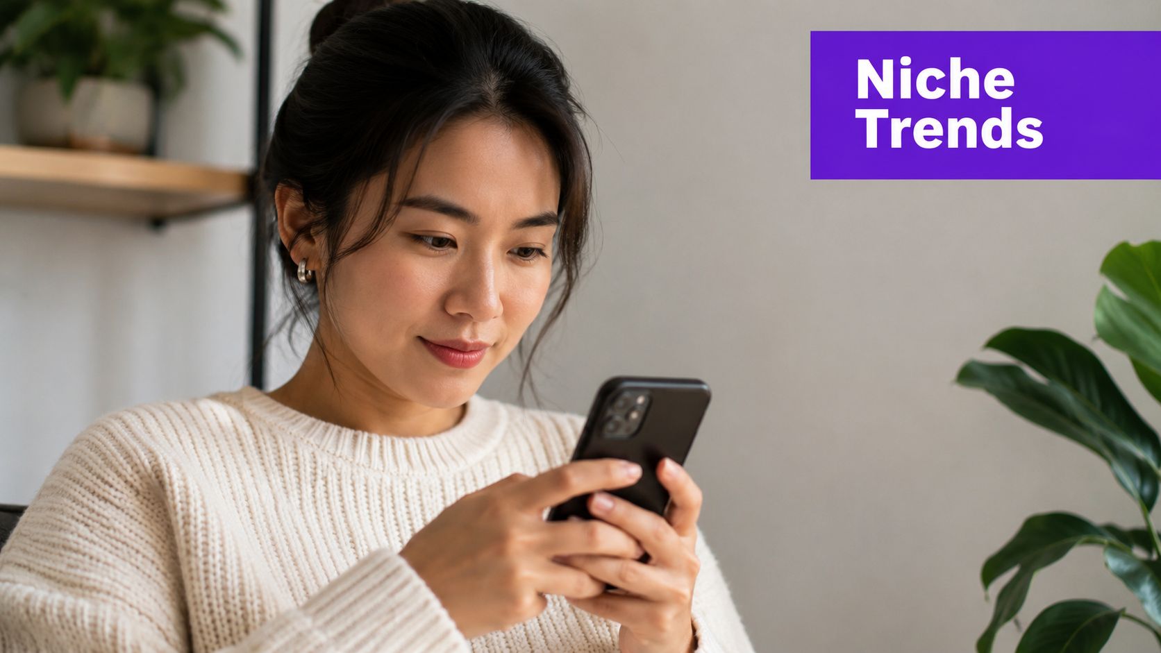 An Asian woman wearing a cream sweater smiling while looking at her smartphone in a bright room.