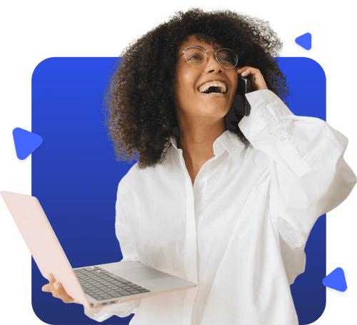 Smiling woman with curly hair and glasses holding a laptop and talking on a phone against a blue background.