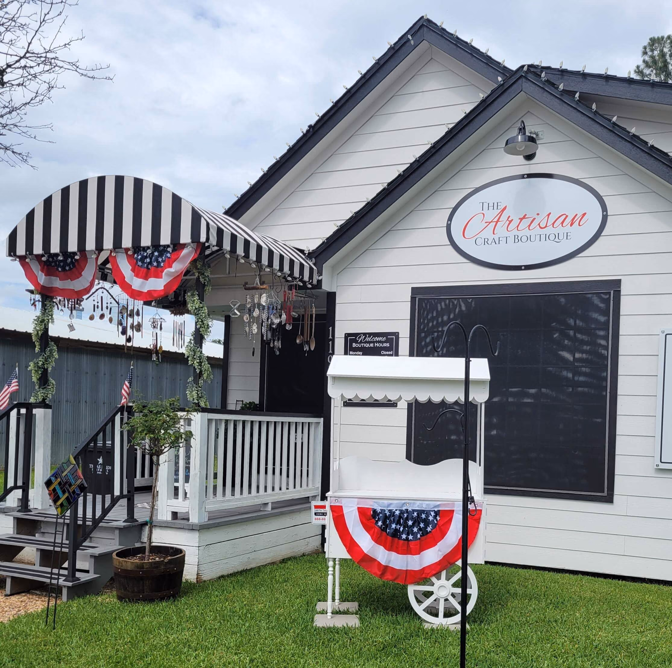 Small white craft boutique with striped awnings, patriotic bunting, and hanging decorations on porch.