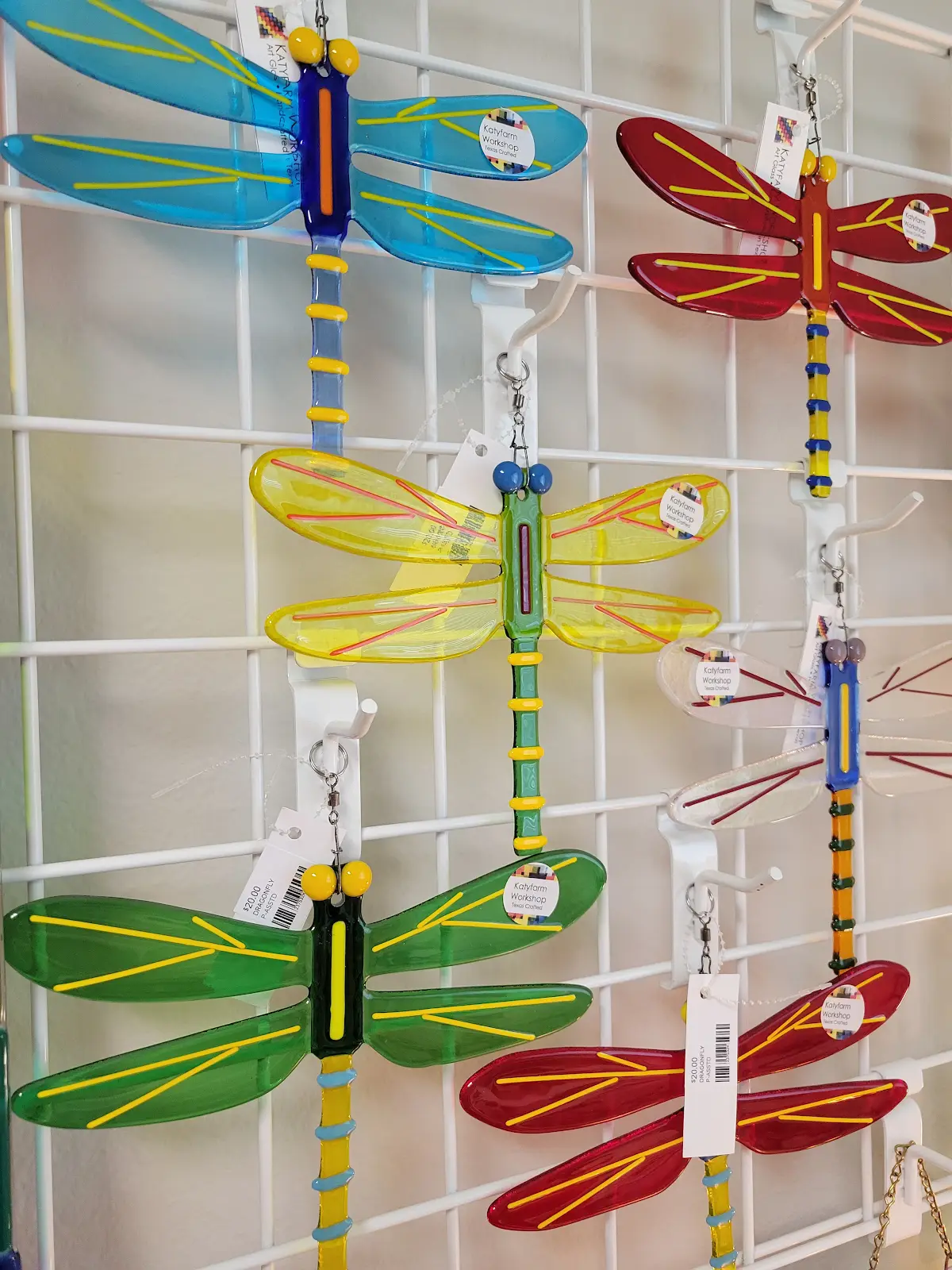 Colorful glass dragonfly ornaments in blue, yellow, green, red, and clear hanging on a white metal grid.