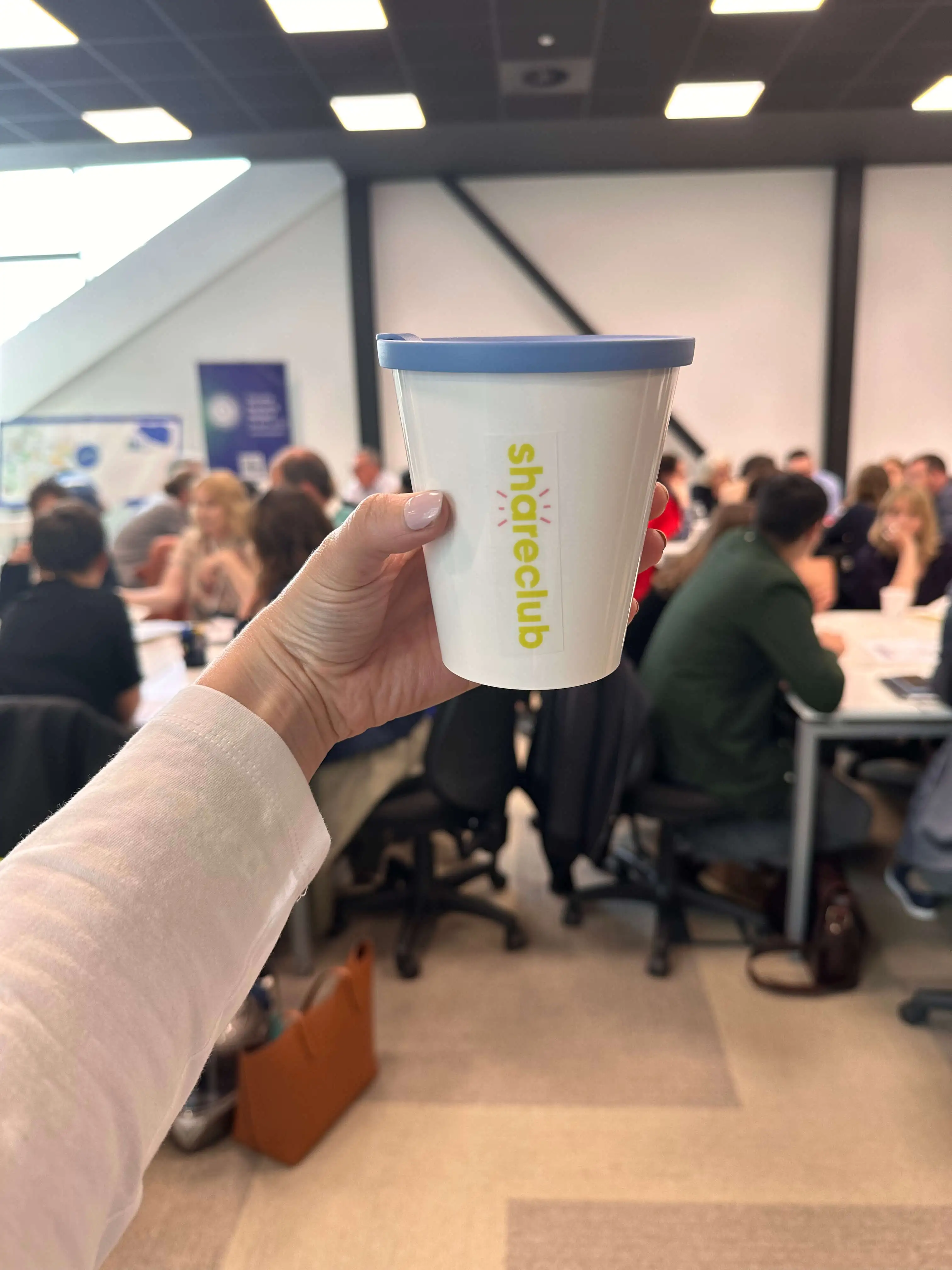A hand is holding a Shareclub cup with the event in the background.